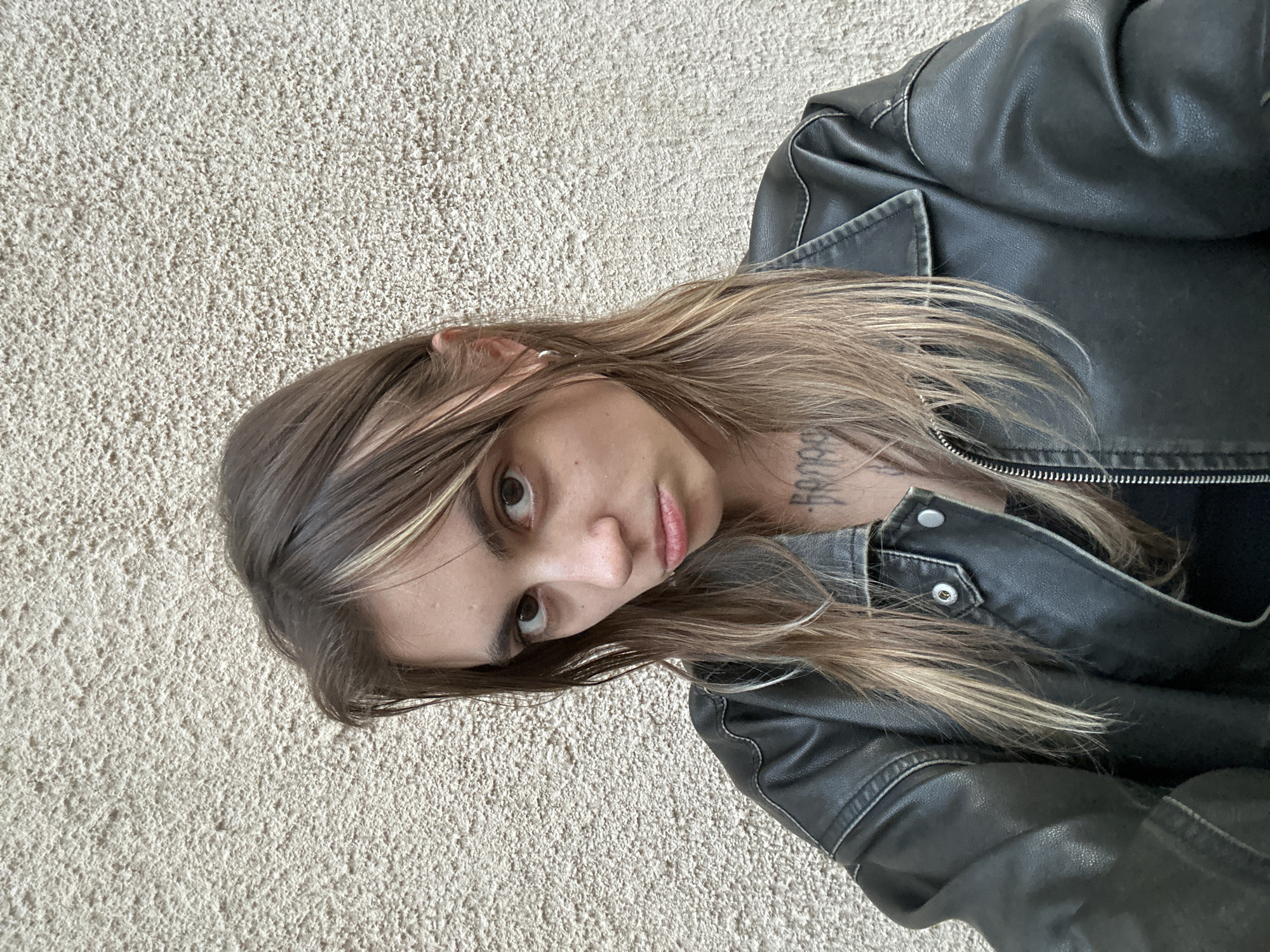 A young Caucasian woman with light brown hair, streaks of blonde, and dark eyes is pictured from the chest up, looking directly at the camera with a pout. She is wearing a black leather jacket over a black top, and has tattoos on her neck and chest. The background is a textured, light-colored wall, possibly stucco, which is common in urban environments. The lighting suggests an outdoor setting during daylight. The provided information about Moscow, Russia, is not visually discernible from the image itself.