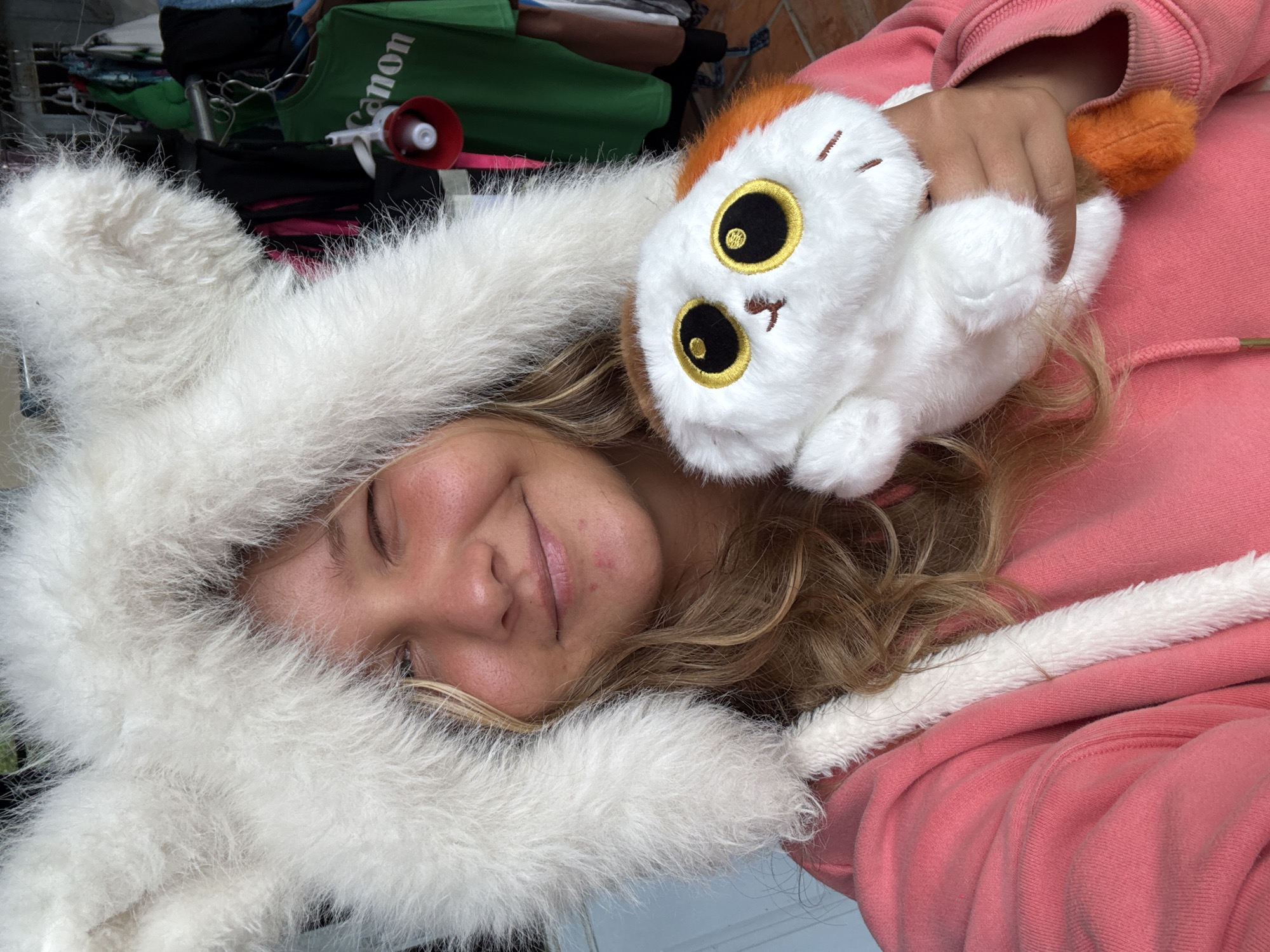 A young woman with blonde hair smiles and winks at the camera while wearing a fluffy white hooded jacket with large ear-like attachments. She is holding a small plush toy with large yellow and black eyes. In the background, clothing items hang on a rack, including a green t-shirt with "Canon" written on it. The scene appears to be outdoors, possibly in a market or shop in the city of Đạ Cháy, Vietnam.