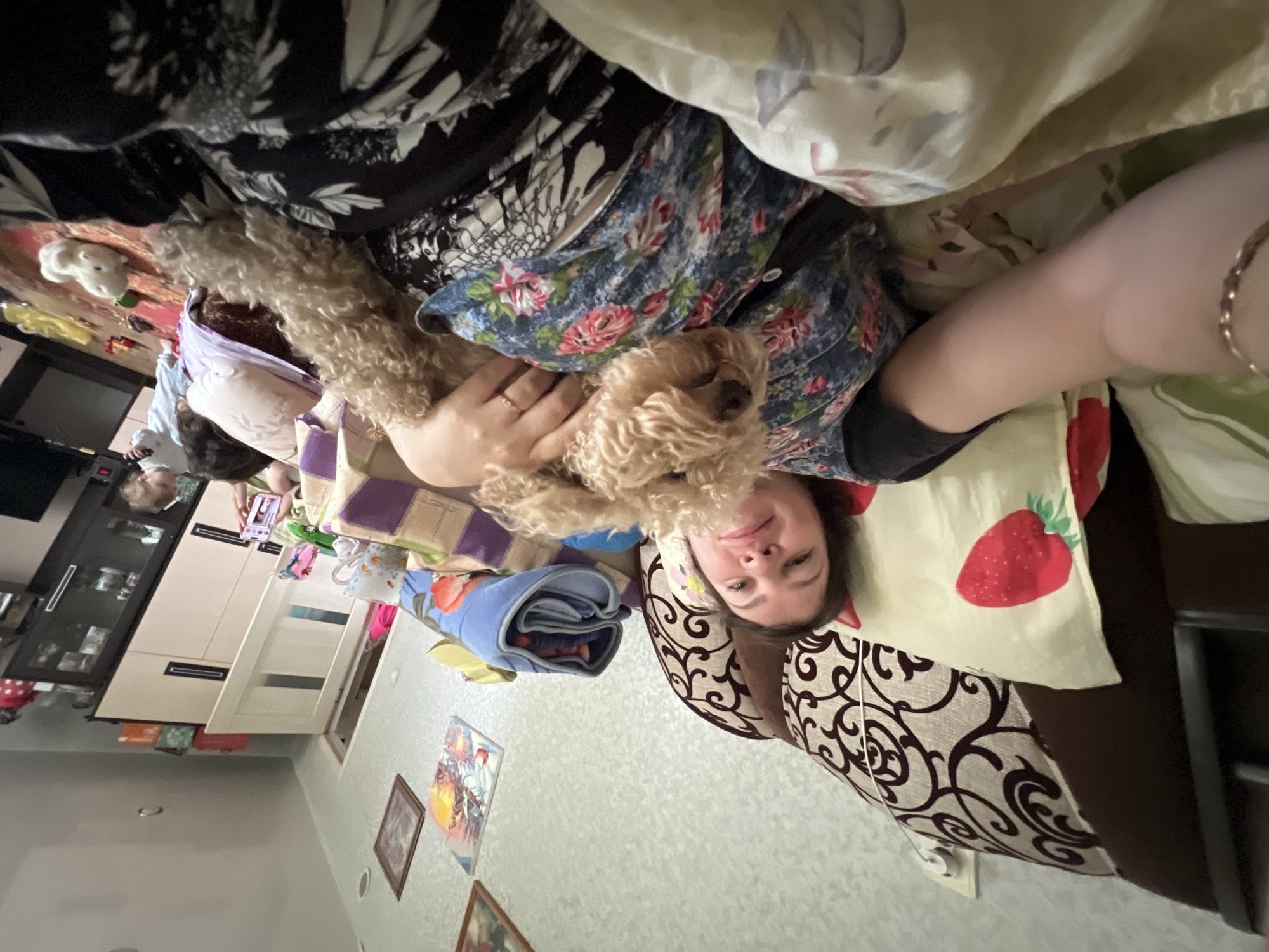 The image shows a woman lying on a bed, holding a light brown poodle. The woman is wearing a dark blue dress with a floral pattern. The poodle is looking at the camera. In the background, a child is visible, playing with toys on the floor. The room appears to be a bedroom, with a bed, pillows, and furniture. The scene is likely located in Panteleevka, Russia, based on the provided context. The overall atmosphere is casual and relaxed.