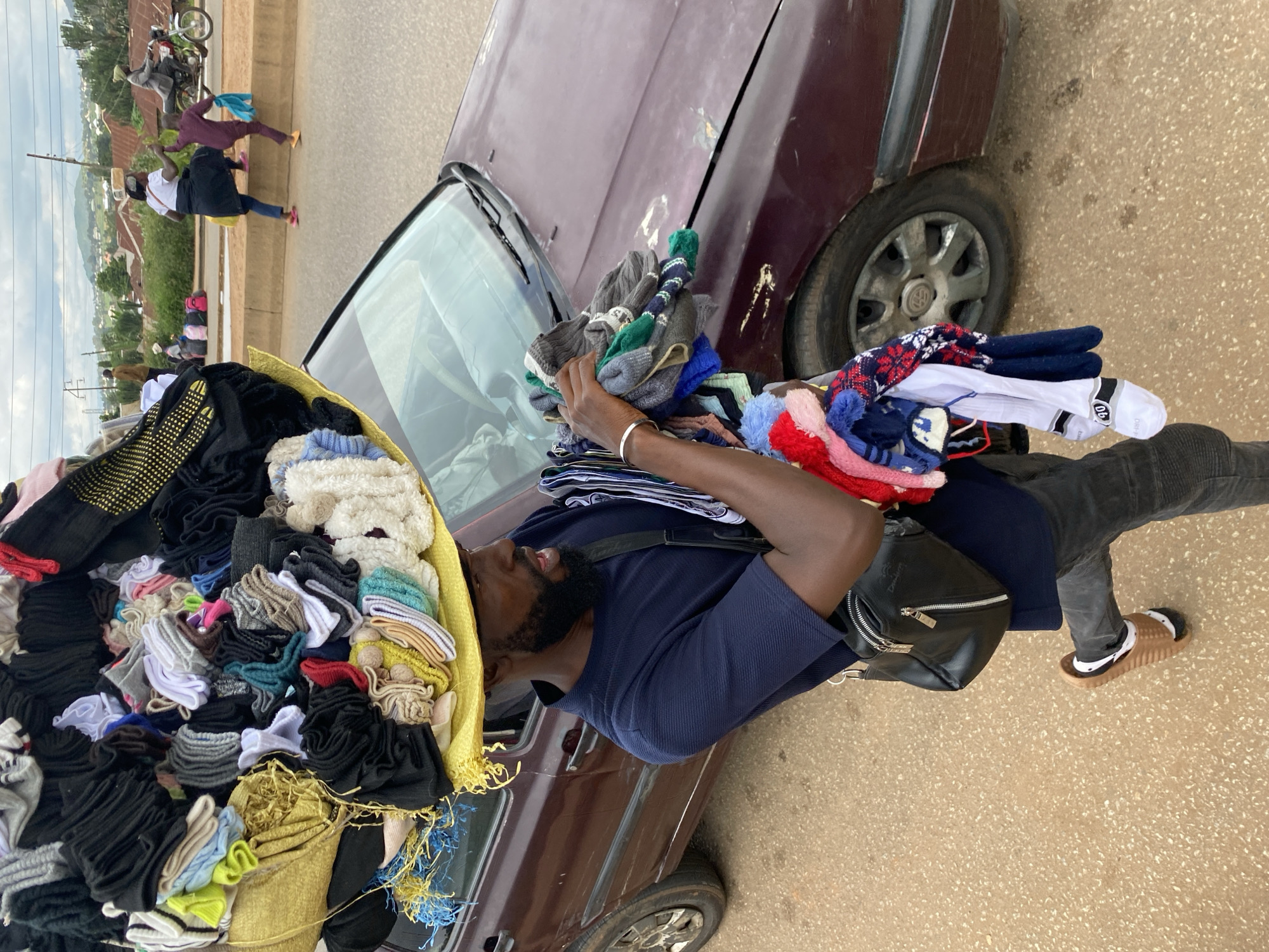 In Bukuru, Nigeria, a man with a beard and a navy blue shirt is walking next to a maroon car. He is carrying a large yellow basket filled with socks and other garments on his head. In his right arm, he holds more socks and a few small knitted hats. He is wearing dark pants and brown sandals with white socks. A black crossbody bag is slung over his left shoulder. In the background, several people are walking on a sidewalk, and a motorcycle is driving on the road. Buildings and trees are visible in the distance, suggesting a residential area. The sky is overcast.