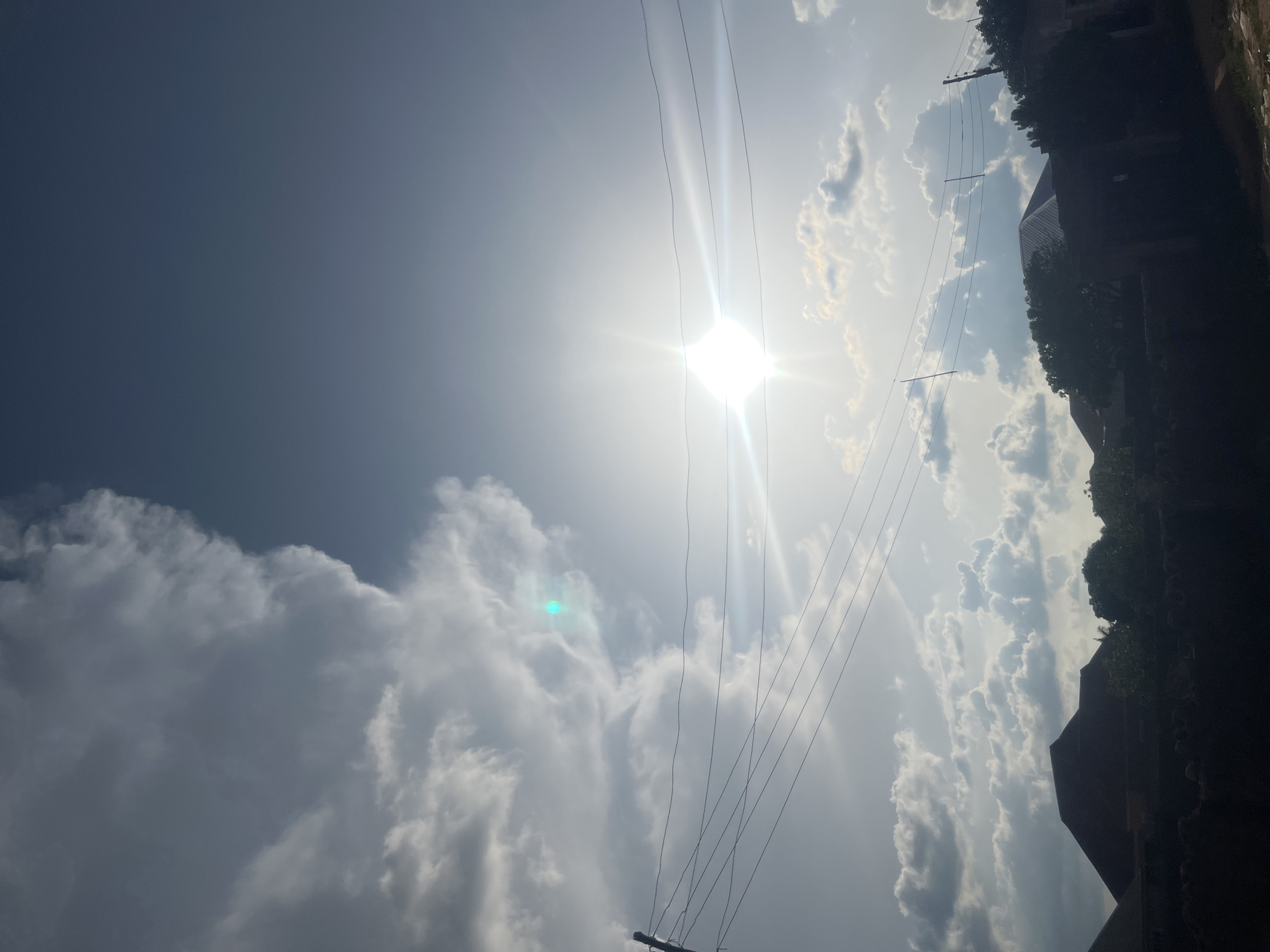 The image depicts an outdoor scene in Barakin Akawo, Nigeria, characterized by a complex sky, utility lines, and the silhouetted rooftops of buildings and trees. The upper left quadrant of the sky is dominated by a dense, dark grey cumulus cloud formation. Contrarily, the middle and right sections of the sky display brighter, scattered white cumulus clouds against a visible blue sky, particularly in the upper right. The sun is a prominent, intensely bright white light source positioned in the middle-right, emitting a strong lens flare and distinct light rays. A notable, circular cyan-green lens flare or reflection is present within the cloud mass in the upper left-center.
Several dark utility wires, likely power or communication lines, stretch horizontally across the mid-ground of the image. These wires originate from a utility pole partially visible on the far left and extend towards another pole on the far right, which is also partially visible.
The lower third of the image consists of the dark, undifferentiated silhouettes of residential structures and dense vegetation. Identifiable architectural elements include a prominent, dark, pyramid-shaped roof in the lower left and a lighter, possibly corrugated metal roof further back and to the right. The silhouettes of numerous trees and bushes are interspersed among these structures, obscuring the ground level. No human subjects or specific actions are discernible within the frame. The overall environment suggests a residential area with common infrastructure and local architectural characteristics.