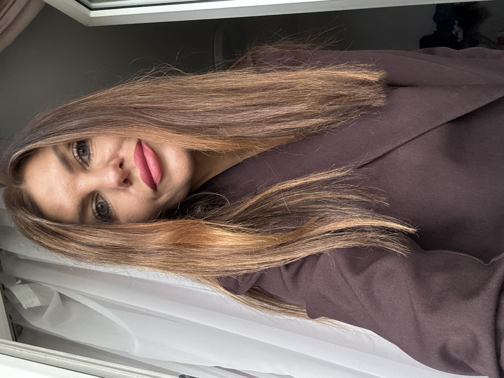 A woman with shoulder-length brown and blonde hair and pink lipstick is seen in an indoor setting. She is wearing a brown collared shirt. In the background is a window with white curtains. The location is in or around Chaikovsky, Russia.