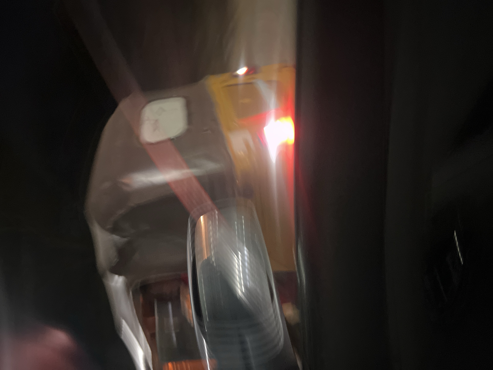 The image captures a blurry, low-light scene, likely at night, characterized by significant motion. The primary subject appears to be a section of a yellow vehicle, possibly a commercial tricycle known as a Keke NAPEP, commonly found in Abuja, Nigeria. A prominent, illuminated red light, consistent with a taillight or brake light, is visible on the right side of the yellow vehicle, with a smaller white light above it.
To the left, a white rectangular object with faint, illegible markings or text is attached to the yellow vehicle, partially obscured by a red strap or band that runs diagonally across it. In the foreground, out of focus, is a clear plastic object, which appears to be a stack of disposable cups. The dark areas on the left and right edges suggest the interior of another vehicle from which the photograph was taken, or the surrounding dark environment. The overall blur indicates motion, suggesting the image was captured while either the subject vehicle or the camera itself was moving. There is no discernible weather information or specific emotional context. No readable text is visible.