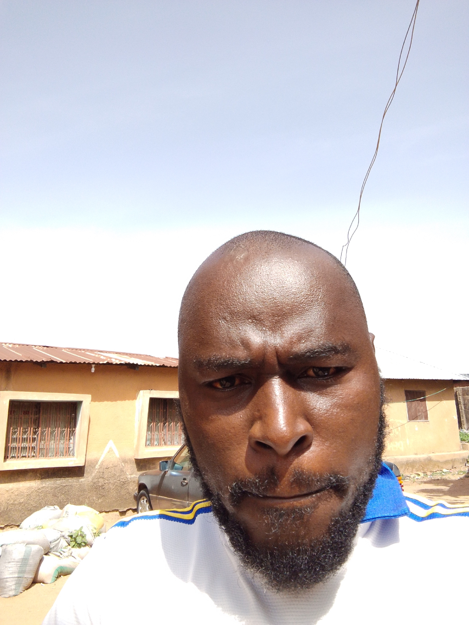 A close-up selfie features a man with dark skin, a shaved head, and a full black beard, looking directly at the camera with a serious, slightly furrowed expression. He wears a white t-shirt with blue and yellow stripes on the collar or shoulder.
The background is an outdoor scene under a bright, clear sky, indicating daytime, likely midday due to the harsh light. On the left side of the frame, several white sacks, possibly containing produce or goods, are stacked on the dusty ground, with some green leaves visible on top of one. Behind the man and the sacks, a light brown building with a rusted corrugated metal roof and windows secured with metal bars is visible. A dark-colored car is partially parked in front of this building. Another similar building can be seen partially on the right. A thin, dark cable or wire extends diagonally from the top right of the frame against the bright sky. The setting appears to be a street or open area in a residential or semi-urban environment, consistent with Jos, Nigeria. There is no visible text in the image.
