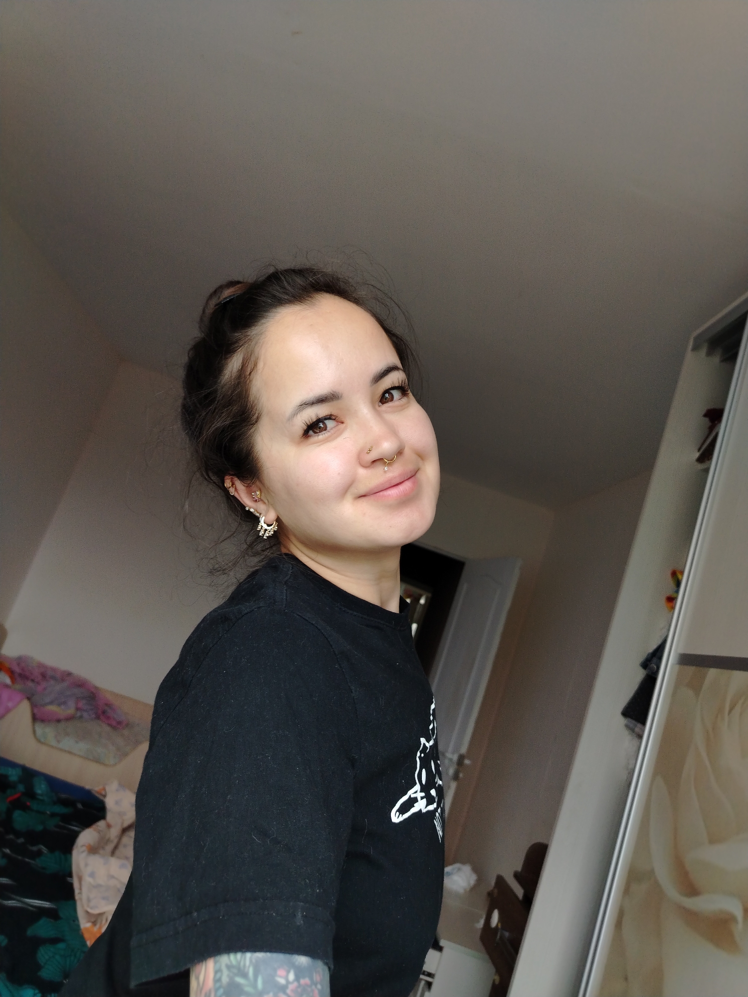 A young woman with brown hair pulled up in a bun and brown eyes is looking to her right and smiling. She is wearing a black t-shirt with a white graphic. She has a nose ring and multiple earrings. She is in a room with a closet and a bed in the background. The image is taken from a slightly high angle.