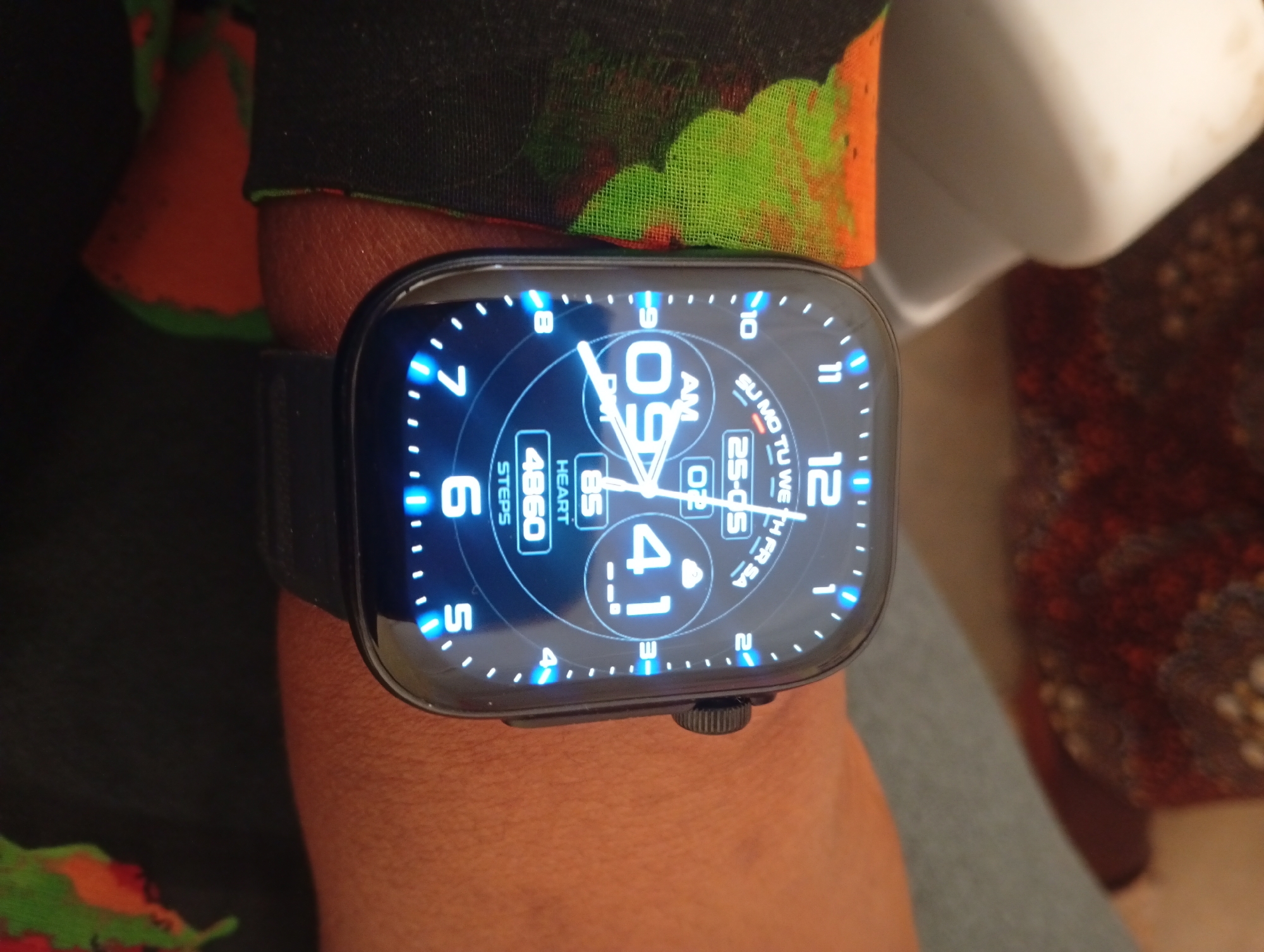 A close-up shot focuses on a wrist-worn smartwatch, showcasing its digital display. The screen prominently features the time as "09:41 AM," along with additional metrics: "4860 Steps" and a heart rate of "85." The watch band appears to be made of a dark, smooth material. The background is partially obscured, with a fabric of mixed colors (orange, green, black) visible in the upper left, and a muted brown pattern appearing in the upper right. The wrist itself, displaying an orange skin tone, provides the immediate context for the watch.