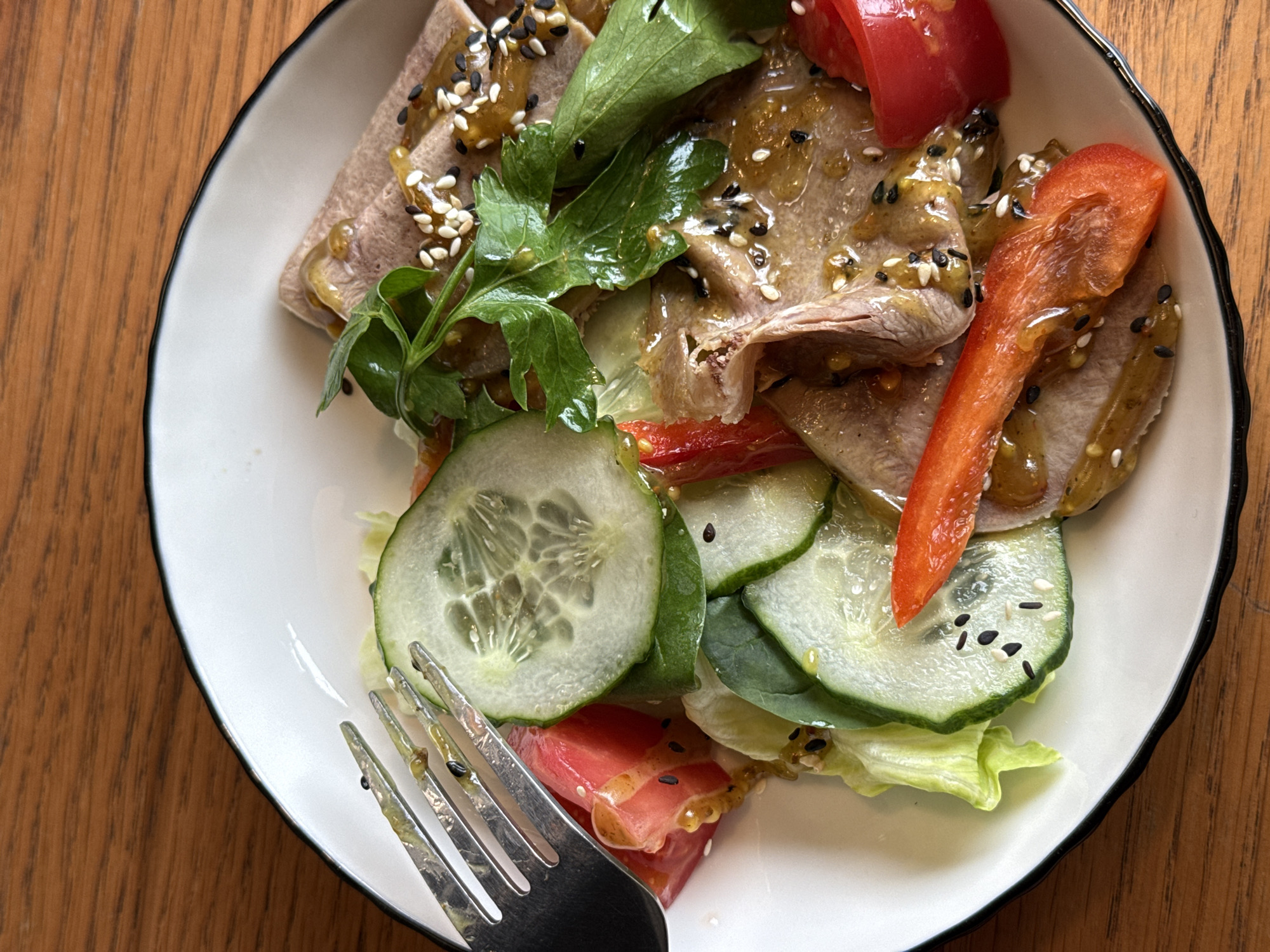 An overhead, slightly angled shot displays a colorful salad served on a white plate with a dark rim, resting on a wooden table in Votkinsk, Russia. The main subject is a salad composed of thinly sliced, cooked meat, likely beef tongue, which is light brown in color. The meat is generously drizzled with a yellowish, somewhat translucent dressing, garnished with both white and black sesame seeds.
The salad also features fresh green components including crisp lettuce leaves, several slices of cucumber, and sprigs of green herbs, possibly parsley or cilantro, adding a vibrant touch. Red elements include chunky pieces of tomato and elongated strips of red bell pepper, providing a pop of color and freshness. A silver fork, with some dressing residue on its tines, is positioned at the bottom left of the plate, suggesting the meal is either about to be consumed or has just begun. The wooden table serves as a warm, natural background, contrasting with the bright white of the plate. There is no visible text in the image.