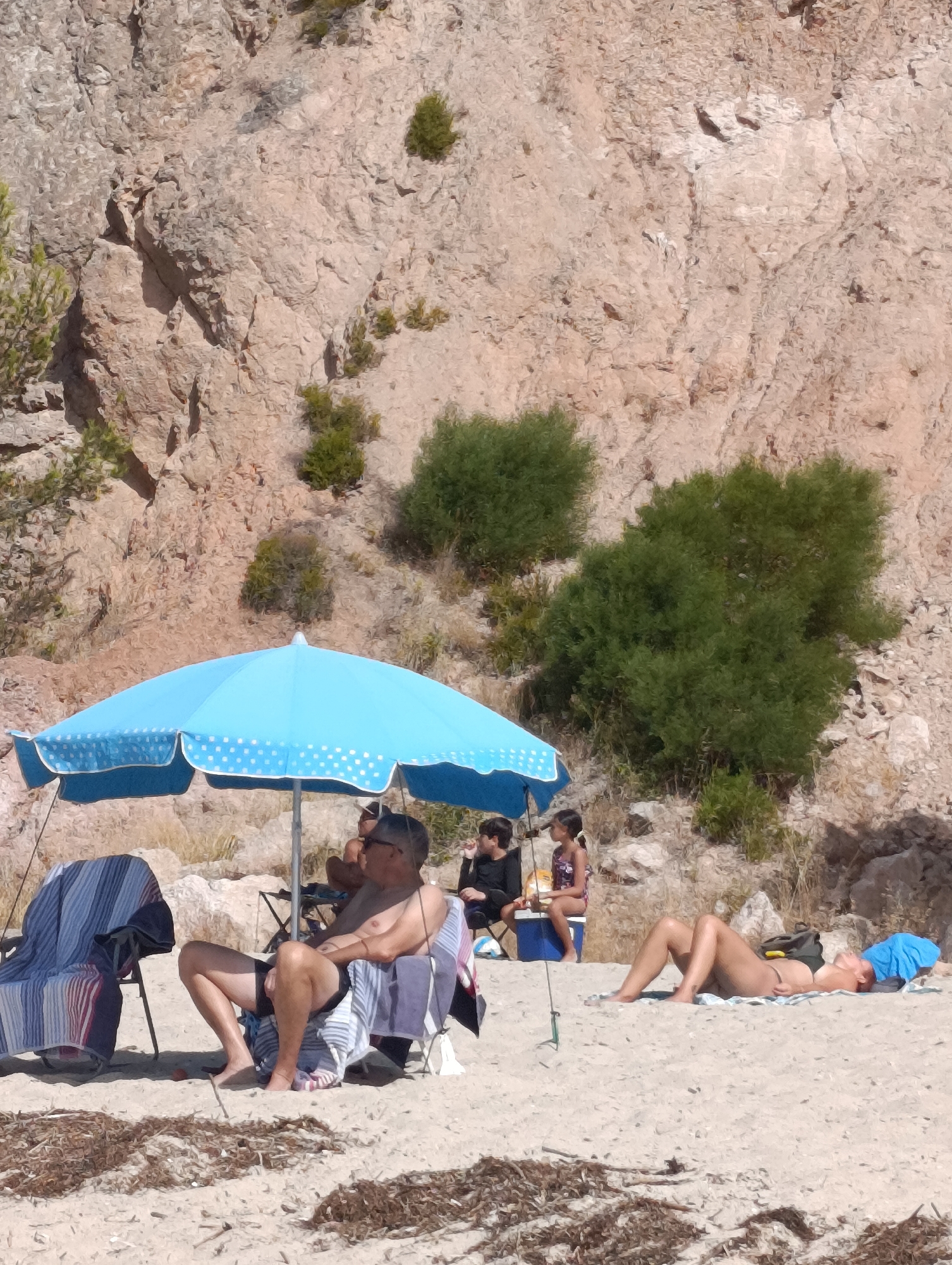 The image depicts a sandy beach with a rocky cliff face in the background. Several people are present. In the foreground, a man sits in a beach chair under a blue and white polka-dotted umbrella. He is wearing black swim trunks and has a towel draped over his chair. To his left, another empty beach chair is covered with striped towels. Further back, a group of people, including two children, are seated on the sand. To the right of the man in the chair, a woman is lying on the sand on a towel, wearing a bikini and a blue headscarf. There are also some pieces of dried seaweed or vegetation scattered on the sand. The scene appears to be a sunny day at the beach. This location could be part of the Arrábida Natural Park, known for its stunning coastline and cliffs near Setúbal, Portugal.