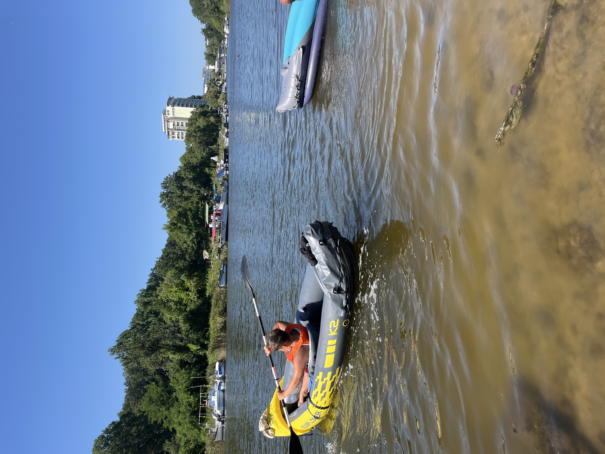 A young person wearing an orange life vest is paddling a yellow and gray inflatable kayak on a body of water. The kayak has "EXPLORER K2" printed on its side. In the background, there are several boats docked along a shoreline lined with trees. A tall, light-colored building is visible among the trees in the distance. The water is murky and appears shallow near the foreground. The sky is clear and blue. This scene is likely located in Primorsko, Bulgaria, given the presence of coastal vegetation, boats, and multi-story buildings indicative of a seaside town.