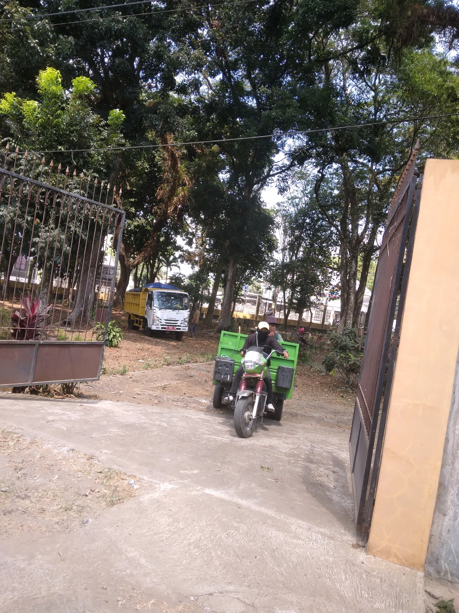The image captures a scene at what appears to be an entrance to a property in Desa Pamijen, Kecamatan Sokaraja, Indonesia. In the foreground, a motorcycle with a green cargo box is being ridden by two individuals. The driver, wearing a white cap and a dark face covering, is seated at the front, while another person is behind them, partially obscured. The motorcycle is on a paved surface.
In the middle ground, a yellow and white truck is parked. A person in a yellow shirt is standing near the truck. The truck has its bed covered with a blue tarp. The area surrounding the truck is unpaved and covered with dry leaves and dirt.
The background features a dense canopy of large trees, suggesting a natural or park-like setting. A metal gate with intricate designs is visible on the left side of the image, partially open. On the right side, a large concrete pillar with a textured surface stands next to another metal gate. The overall lighting indicates it is daytime, with sunlight filtering through the trees. The scene seems to depict a moment of activity related to transportation and possibly work, given the presence of the truck and the cargo on the motorcycle.