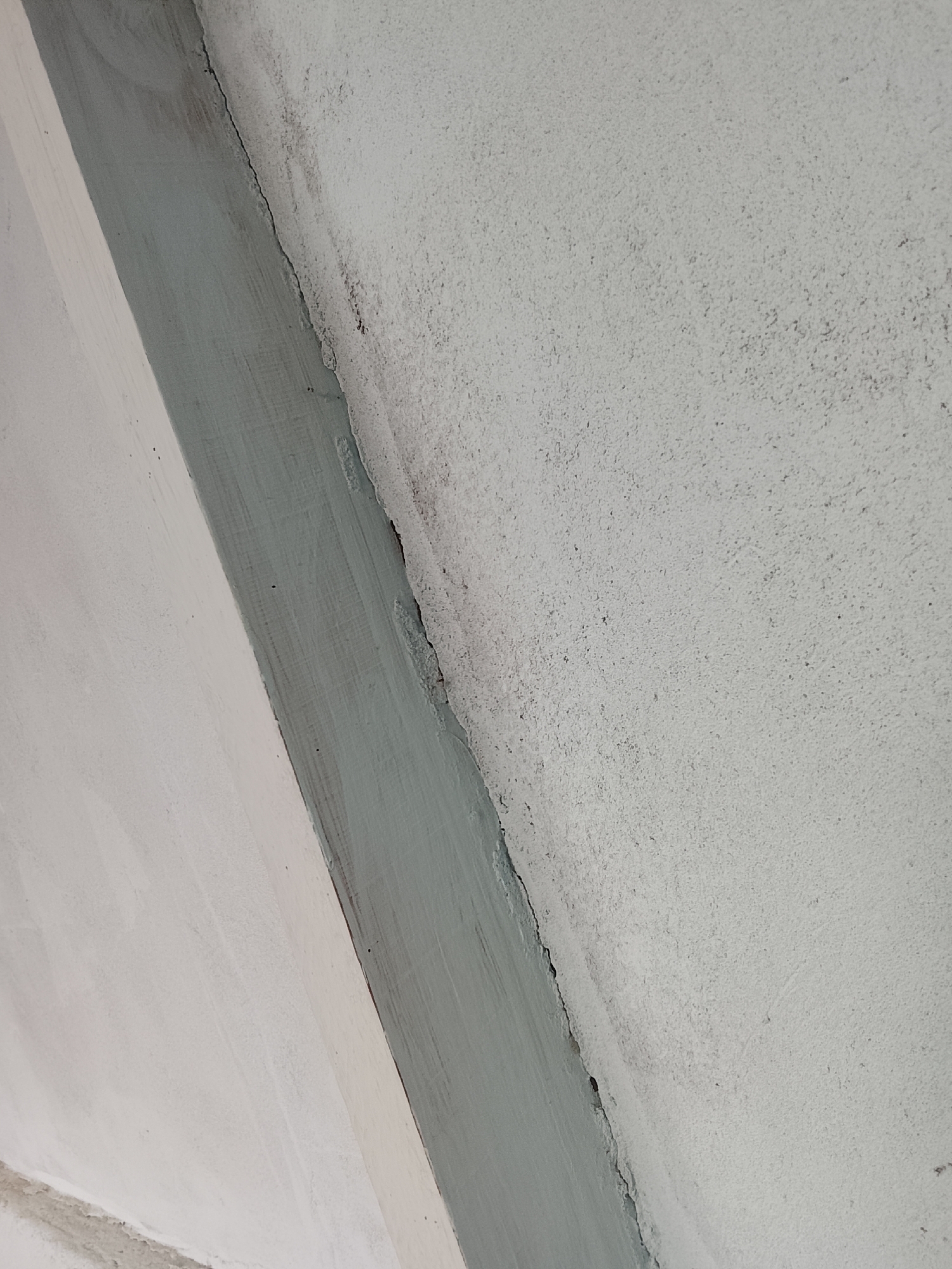 The image shows a close-up, angled view of a textured, light-colored wall with a green-painted strip running diagonally across it. A thin, dark crack is visible along the edge where the painted strip meets the wall. The wall surface is rough and speckled with small dark particles, suggesting it is made of concrete or stucco. There are no people or specific landmarks visible to indicate a precise location within Mehnatabad, Tajikistan. The scene appears to be an interior or exterior wall detail.