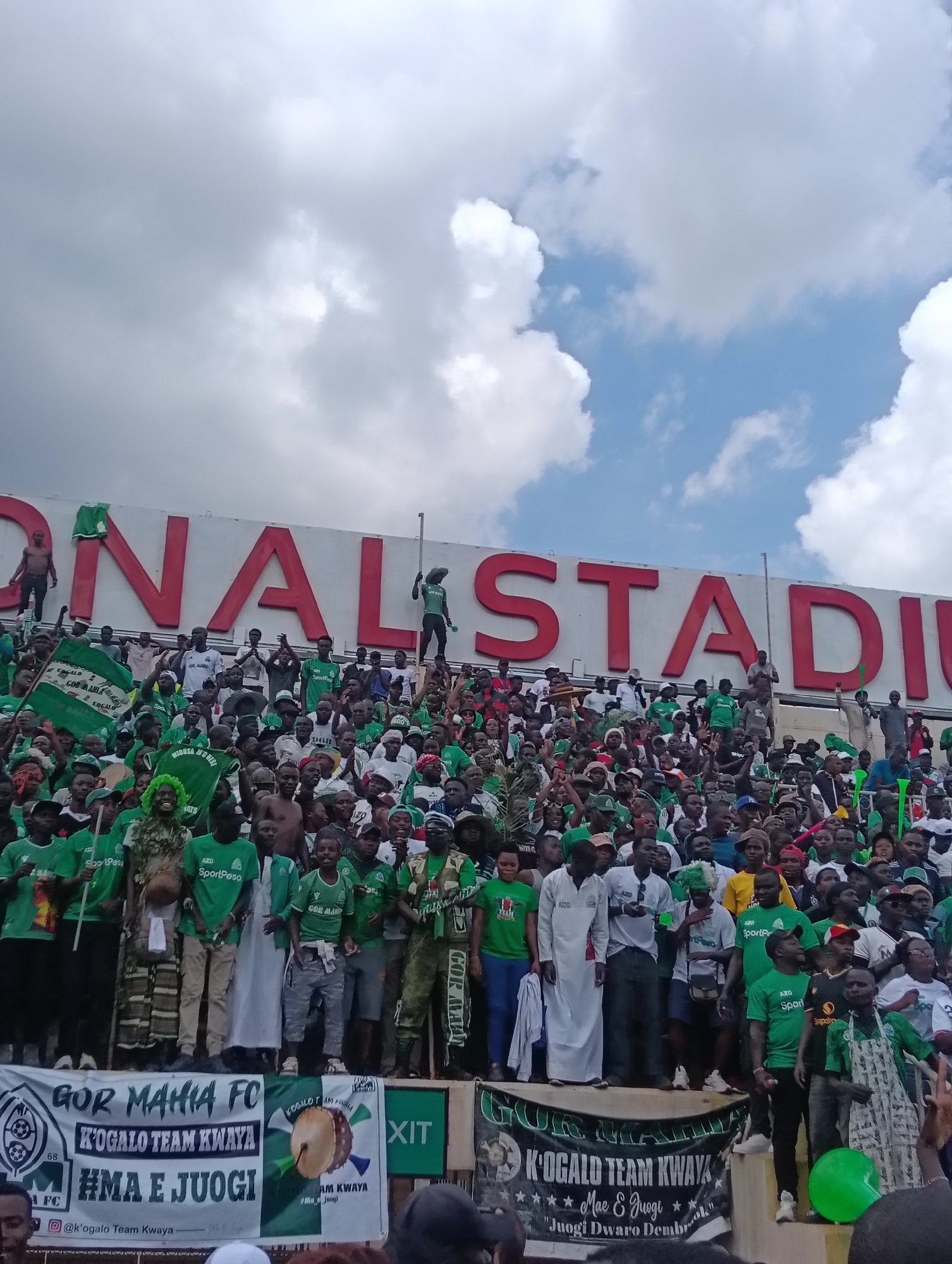 The image shows a large crowd of people, predominantly wearing green and white, gathered at a stadium. The stadium's name, "NATIONAL STADIUM," is prominently displayed in red letters. Many people are waving flags or holding up green objects. A banner at the front reads "GOR MAHIA FC" and "K'OGALO TEAM KWAYA" and "HMA E JUOGI". Given the team's name, the colors, and the name of the stadium, this likely depicts a sporting event related to the Gor Mahia football club at the Moi International Sports Centre in Nairobi, Kenya. Several people are standing on an elevated platform in front of the crowd, interacting with the fans. The sky is partly cloudy.