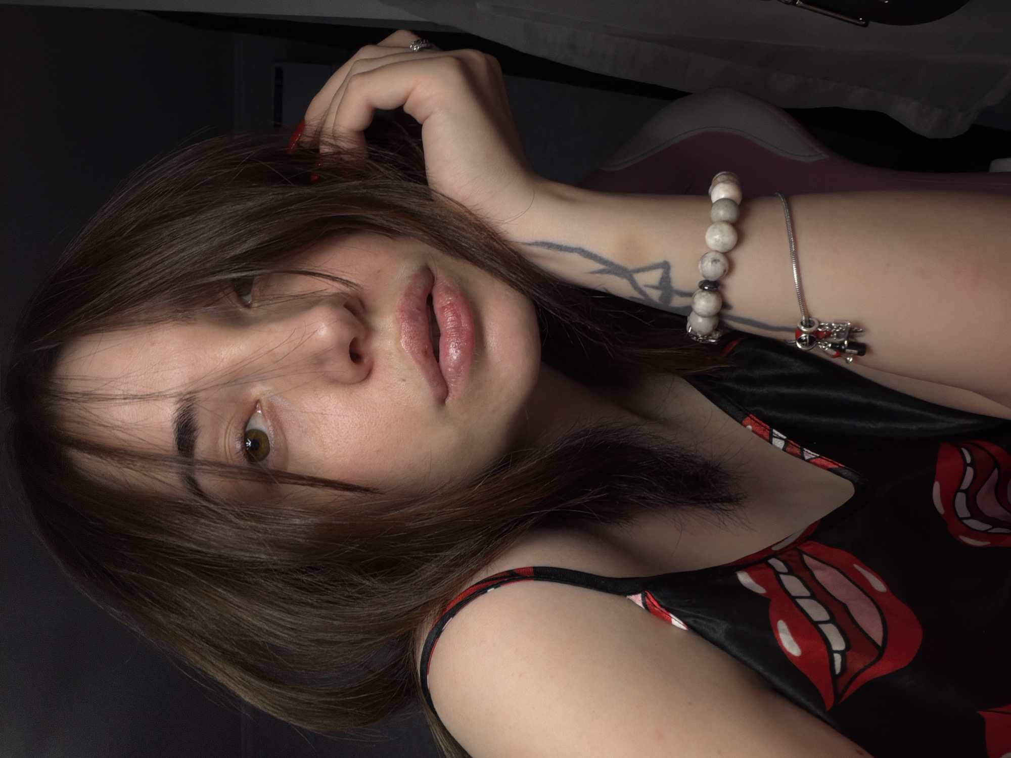 A young woman with brown hair and light skin is shown in a close-up, candid portrait. She is wearing a black camisole with a bold red and white lips pattern. Her left arm is resting on her hand, which is supporting her face. She has a tattoo on her left forearm and is wearing a bracelet with light-colored beads and a charm. Her lips are slightly parted, and her eyes are looking towards the camera. The background is dark and nondescript, suggesting an indoor setting. The lighting is soft, highlighting her face and the details of her clothing and accessories.