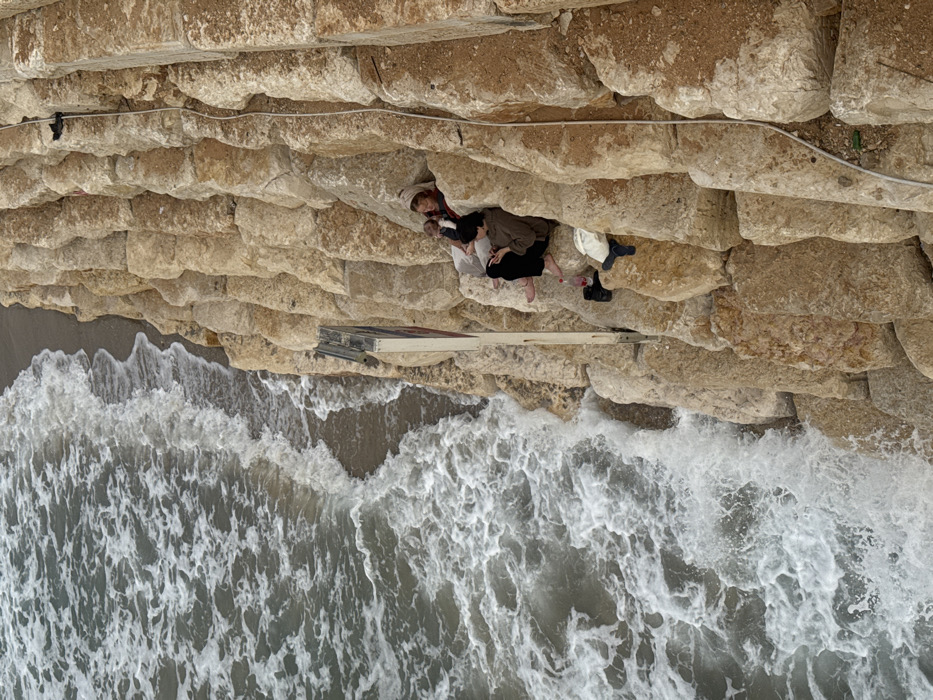 The image displays a high-angle view of a section of the Tel Aviv-Jaffa coastline in Israel, characterized by a steep, terraced sea cliff composed of large, light-colored stone blocks. To the left, the Mediterranean Sea features choppy water with white-capped waves breaking onto a narrow strip of dark sand at the base of the cliff.
Two individuals are positioned on a recessed ledge within the cliff face, approximately midway down its visible height. The individual on the left is dressed in light-colored clothing, including a head covering or hood, and appears to be holding a bundled object or a small child wrapped in a dark fabric. Their gaze is directed generally towards the right. The individual on the right is seated beside the first, wearing dark pants and a brown long-sleeved top, and is looking down at a handheld device. A red plastic bottle and a pair of dark footwear are visible on the ledge near this individual. A light-colored piece of fabric or bag is situated just below their feet.
To the upper left of the seated individuals, a vertical, light-colored structure, possibly a signpost with dark lettering, is affixed to the cliff face. A white cable runs diagonally downwards from the upper right portion of the cliff, secured at intervals. The overall environment is rugged coastal terrain, with the rock formations showing signs of weathering and construction.
