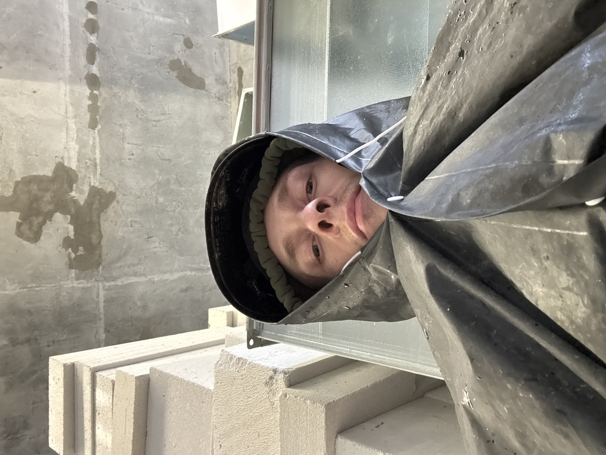 The image shows a man's face from a low-angle perspective, wearing a black hood that is part of a garment. He is wearing a dark hat under the hood, and there's a green elastic band visible around his forehead. The man has light skin and short brown hair. His expression is neutral, with his mouth slightly open. The background consists of a grey concrete ceiling with some stains and imperfections. To the left of the man, there is a stack of light-colored, rectangular blocks, possibly made of concrete or foam. To the right and behind him, there are some metallic structures and a ribbed surface, suggesting an industrial or construction environment. The overall lighting is moderate, and the image appears to be taken indoors. No specific details are provided about the location in Moscow, Russia.