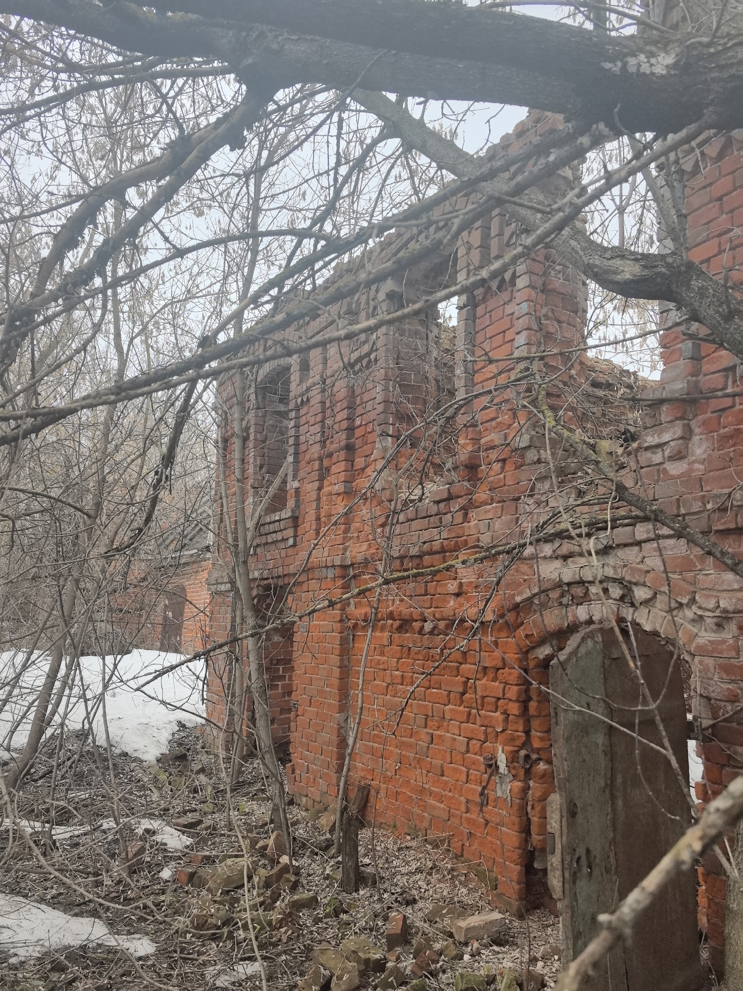 The image shows the ruins of a brick building located in Tolba, Russia. The building is partially collapsed, with sections of the brick walls still standing. There are several window and door openings in the remaining walls. The building is surrounded by bare trees and undergrowth. Patches of snow are visible on the ground. The scene is desolate and suggests a state of abandonment or decay. There are no people visible.