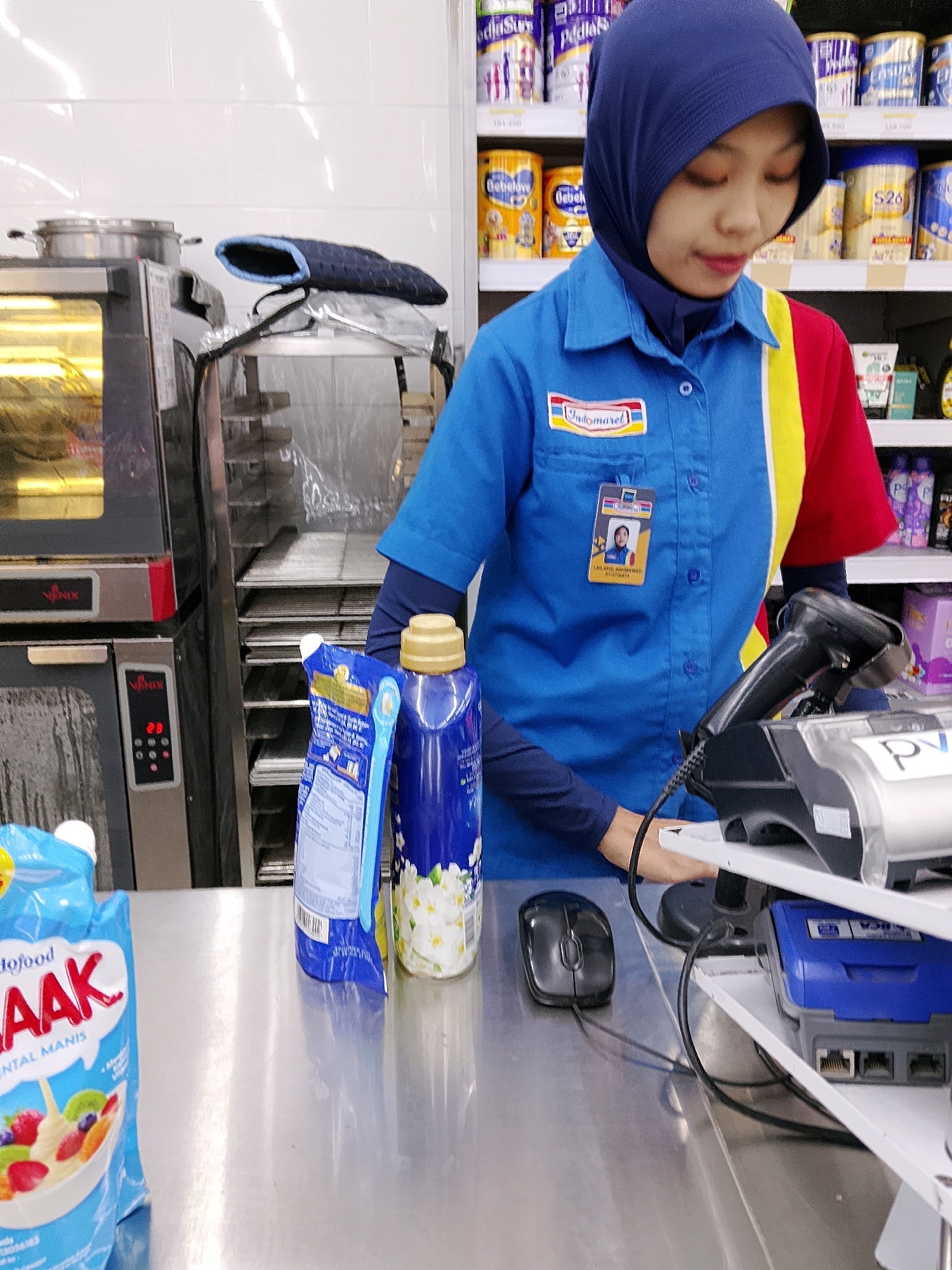 The image shows a young woman wearing a blue and red uniform, a blue hijab, and a name tag. She is standing behind a counter in what appears to be a convenience store in Malang, Indonesia. On the counter, there is a black computer mouse, a barcode scanner, and two cleaning product bottles, one blue with a floral pattern and another with a white pouch. Behind the counter, shelves are stocked with various products. To the left of the woman, there is a commercial oven with a digital display. The overall environment suggests a retail setting, likely a minimarket.