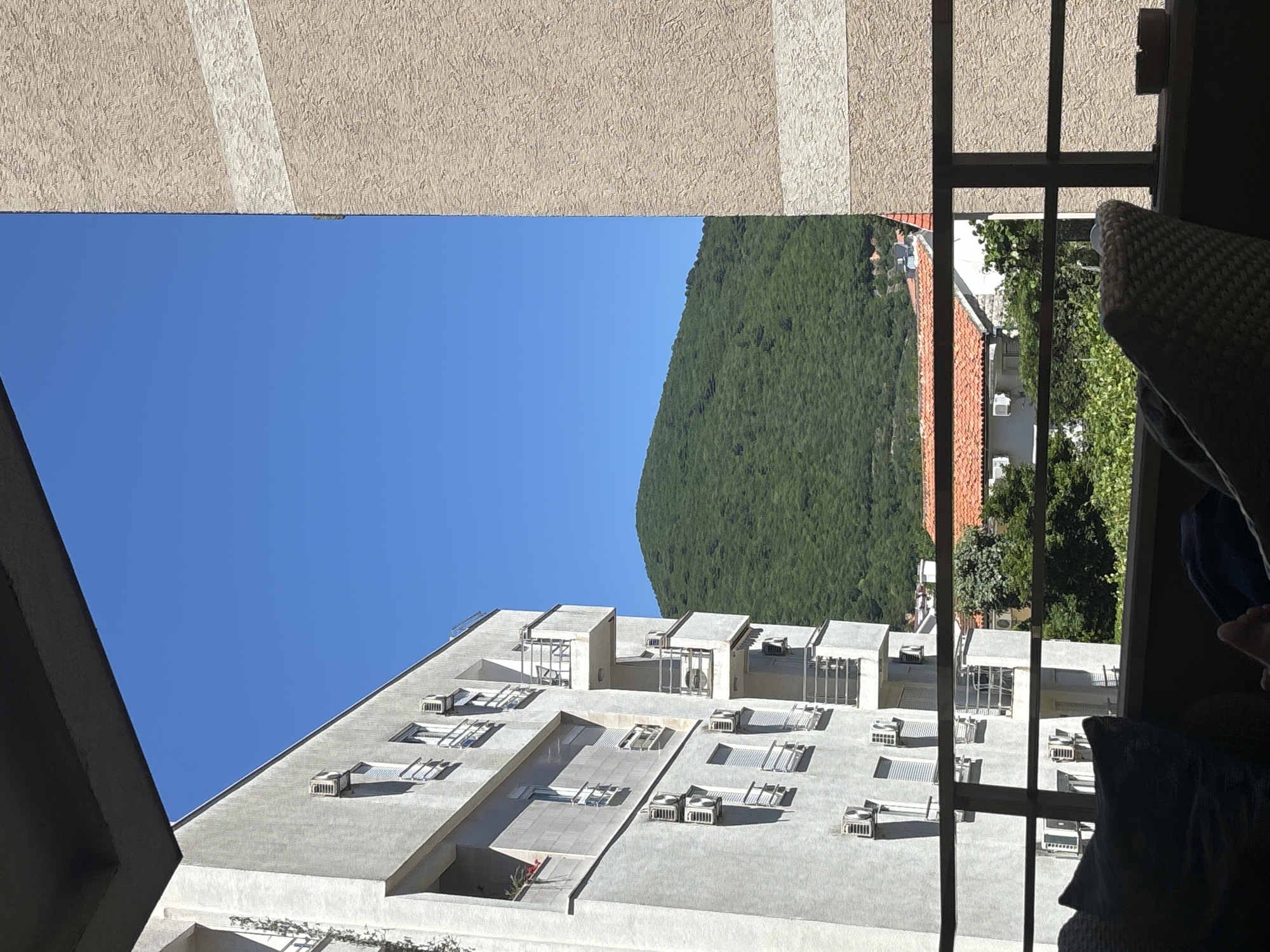 The photograph depicts a view from an interior space, possibly a hotel room, looking out onto an urban landscape in Budva, Montenegro. In the foreground, window frames partially obscure the view. Outside, a multi-story white building dominates the scene, its flat roof dotted with air conditioning units and geometric structures. Behind the building, a lush green mountain rises steeply. A bright blue sky fills the space above the building and mountain. To the right, glimpses of other buildings with red tiled roofs are visible. The interior includes a woven chair and pillows that are partially visible on the right side of the frame. The photograph is taken in daylight, with clear visibility.