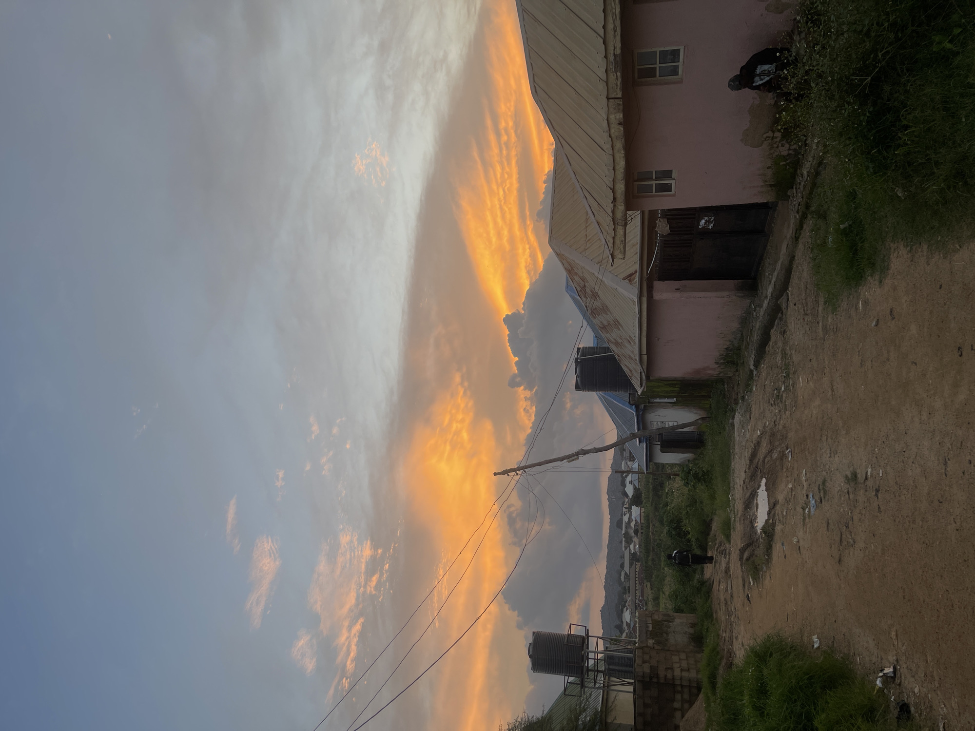 This is a medium shot of a street in Rafin Par, Nigeria, during a sunset. The sky is filled with dramatic orange and yellow clouds, indicative of the setting sun. In the foreground, a dirt road leads into the distance. On the right side of the road, there are two pink buildings with corrugated metal roofs. One building has a large dark gate. A person is sitting in the bushes next to this building. On the left side of the road, there are more buildings, including one with a large black water tank on top. A person is walking away from the camera down the dirt road. Power lines are strung across the scene. The overall impression is of a rural or semi-urban environment.
