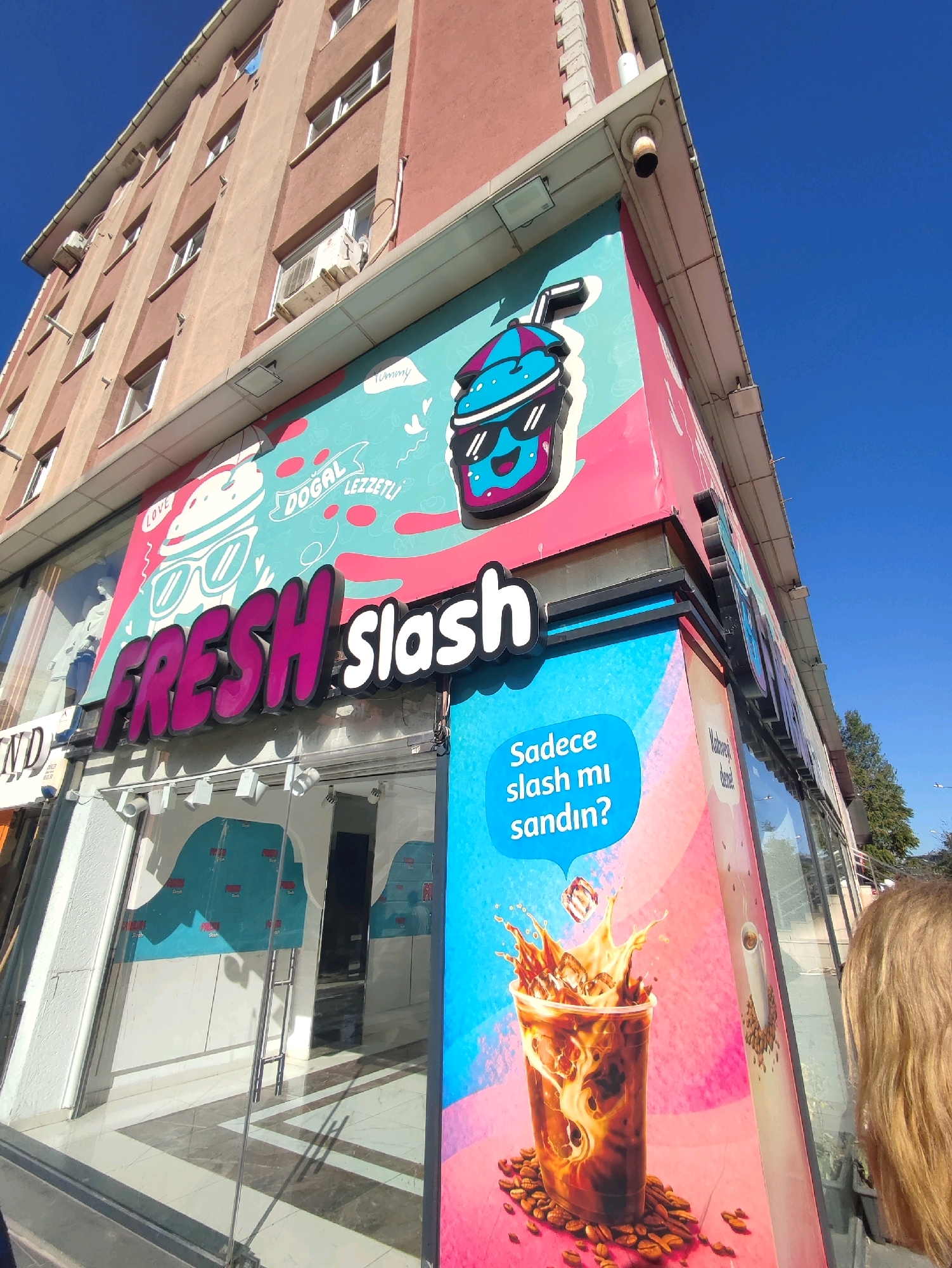 An exterior view of a store with large windows and a colorful facade in Gungoren, Turkey. The building has multiple stories with a salmon-colored exterior and cream-colored trim. Several windows are visible on the upper floors, some with air conditioning units. The store itself features a prominent sign that reads "FRESH slash" in large, pink and white lettering. The facade is decorated with a large graphic of a cartoon character holding a drink and a picture of an iced coffee with coffee beans. Text in Turkish appears on the graphics. A portion of a person's head and shoulder, with blonde hair, is visible on the right side of the frame, suggesting they are looking at the store. The sky above is clear and blue.