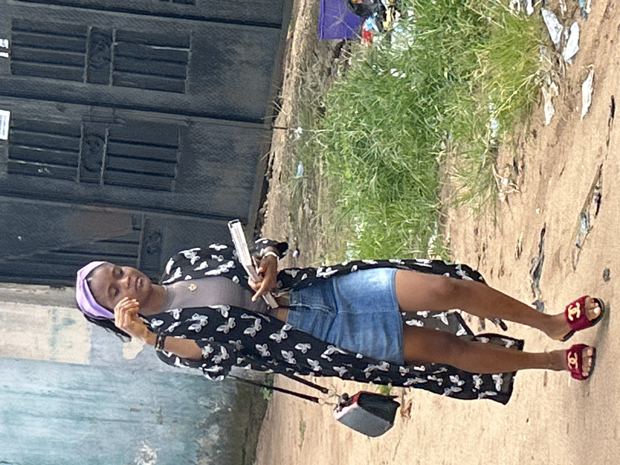 A young woman stands on a dirt path in Ihiagwa, Nigeria. She wears a lilac headband, grey top, denim skirt, and a black butterfly-print kimono. Red sandals adorn her feet. She holds a reflective item and a small purse hangs at her side. A multi-paneled metal door stands to her left, and vegetation borders the path to her right. Debris is scattered on the path's edge.