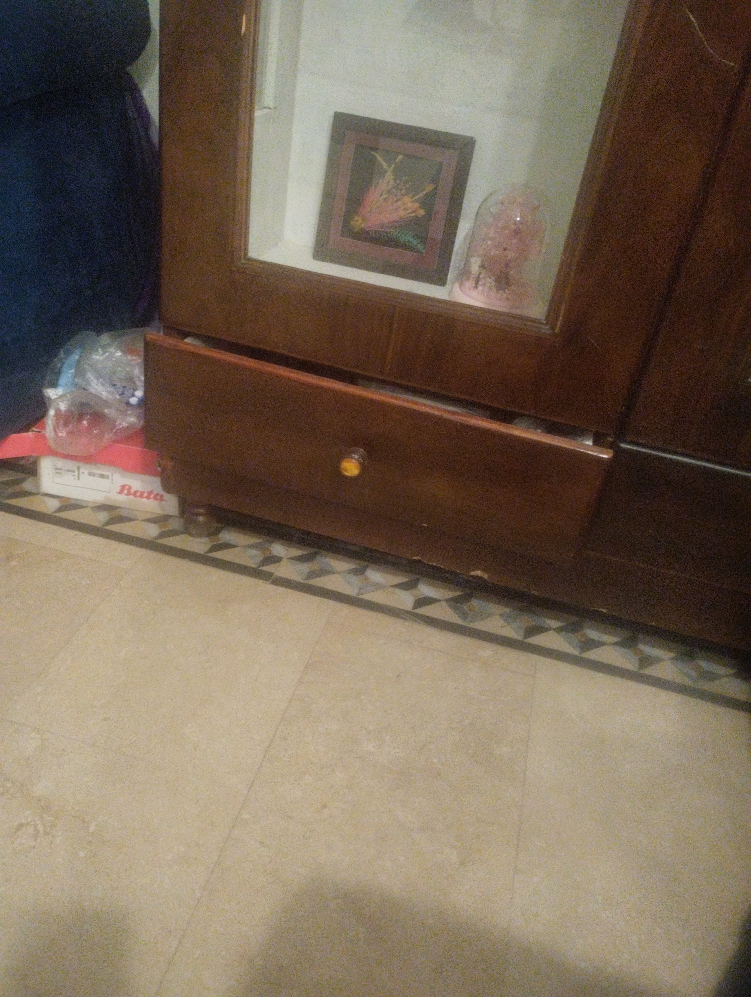 An indoor scene shows a wooden cabinet with a partially opened drawer. Inside the drawer, a red shoe box with "Bata" written on it is visible, along with some plastic-wrapped items. Above the drawer, a glass cabinet displays a framed picture and a glass dome containing pink floral decorations. The floor is tiled with a patterned border. To the left of the cabinet, a dark blue fabric is draped. The scene appears to be in a domestic setting.