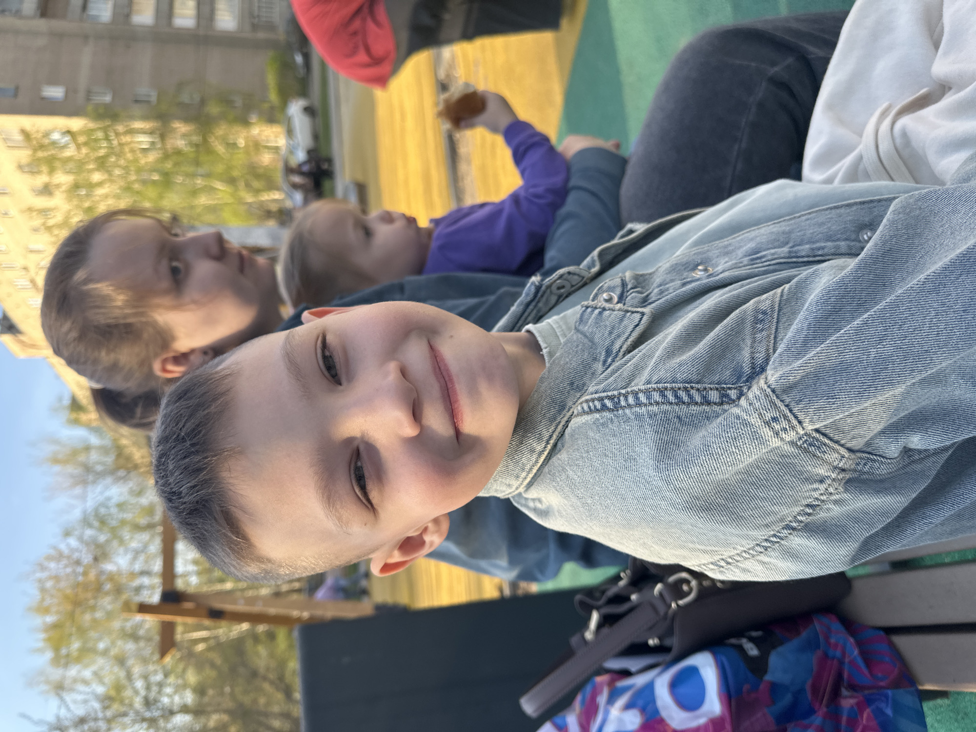 The image depicts a family gathering outdoors, possibly in a park or recreation area in Tomilino, Russia. A child in a light blue denim jacket is the focal point, facing the camera with a slight smile. Behind the child is a woman with her hair pulled back, and a toddler wearing a purple shirt holding food in their hand. The group appears to be sitting or lying on a green surface, likely grass or a picnic blanket. A black purse sits on the ground next to a blue and red patterned bag. The background includes trees and a building in the distance, suggesting an urban or suburban setting.