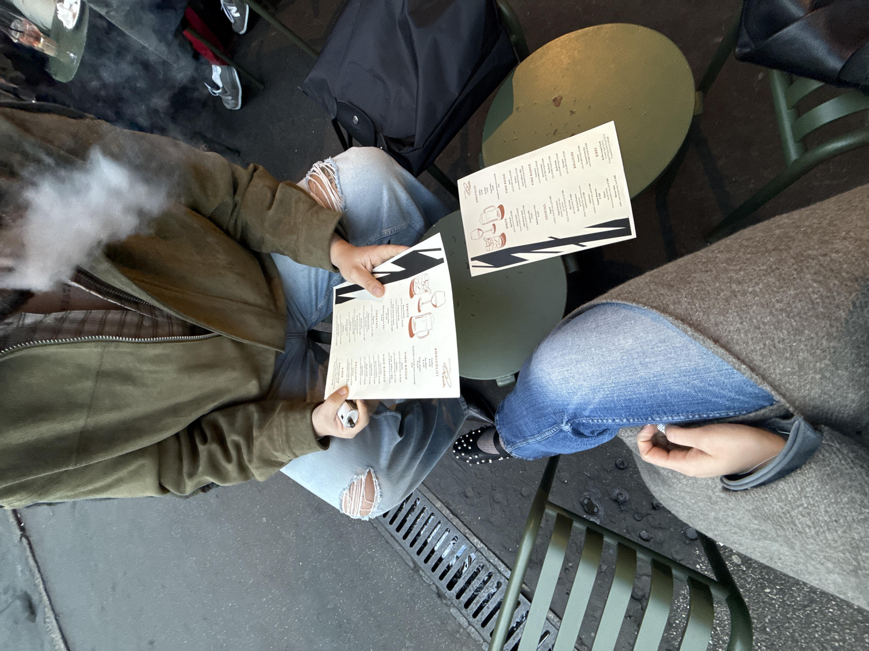 The image displays an outdoor cafe scene from an overhead perspective, likely situated in the Brera district of Milan, Italy, as indicated by "Osteria Brera - Spazio di Milano" visible on the menus.
Two individuals are seated at small, round, dark green metal tables on matching slat chairs. In the foreground, the lower body of one person is visible, wearing light blue jeans, black flat shoes with white studded embellishments, and black socks, partially covered by a greyish-brown woolen garment. A right hand, belonging to this individual, holds a small, light-colored object.
Opposite, a second individual is seen from the chest down. They wear an olive-green zippered jacket over a light-colored plaid shirt and distressed light blue jeans with rips at both knees. This person is holding an open menu with both hands, examining its contents. Their left hand also holds a small white/silver device, from which a cloud of white vapor is being exhaled, obscuring part of their upper chest. A second, identical menu lies flat on the table closer to the foreground. The menus feature Italian beverage categories such as "BIRRE," "VINO BIANCO," "COCKTAIL," and "SPRITZ," with prices listed in Euros. A black foldable tote bag rests on the ground next to this person's chair.
In the background, a partially visible table displays a glass containing a reddish beverage and a small white plate with snacks. The feet of a third person, wearing grey New Balance sneakers, are also visible. The ground surface is dark grey asphalt, with a metal drainage grate positioned near the foreground person's feet.