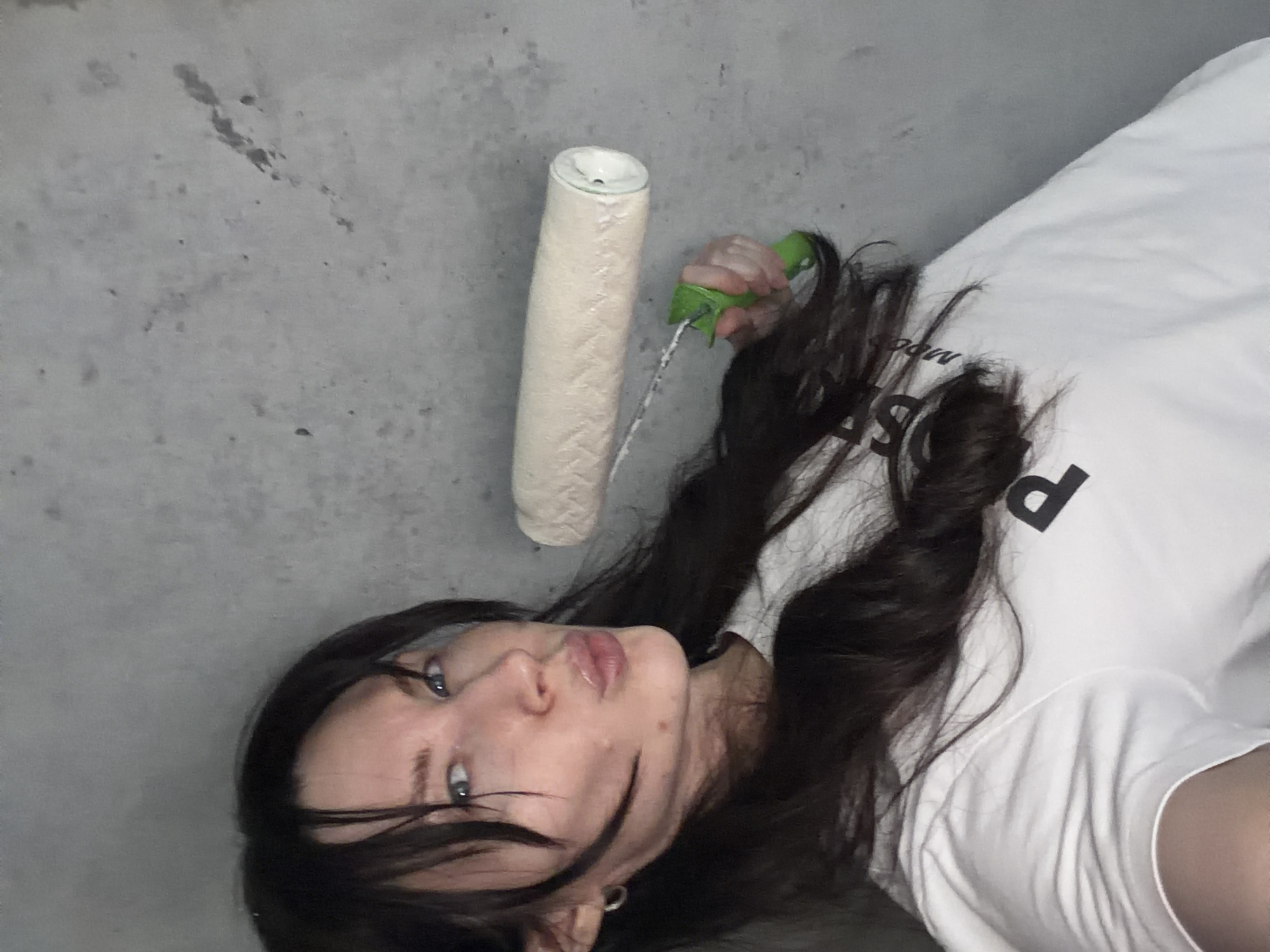 A young adult female is depicted in a medium shot against a light grey, textured wall in Perm, Russia. She possesses long, dark hair, blue eyes, and light skin, and is seen looking directly into the camera with her lips puckered. She wears a white t-shirt featuring a black letter "P" on the chest and a small silver hoop earring in her left ear. Her right hand holds a paint roller with a cream-colored, textured cover and a green handle, extended horizontally towards the right. The background wall shows minor surface imperfections and discoloration.