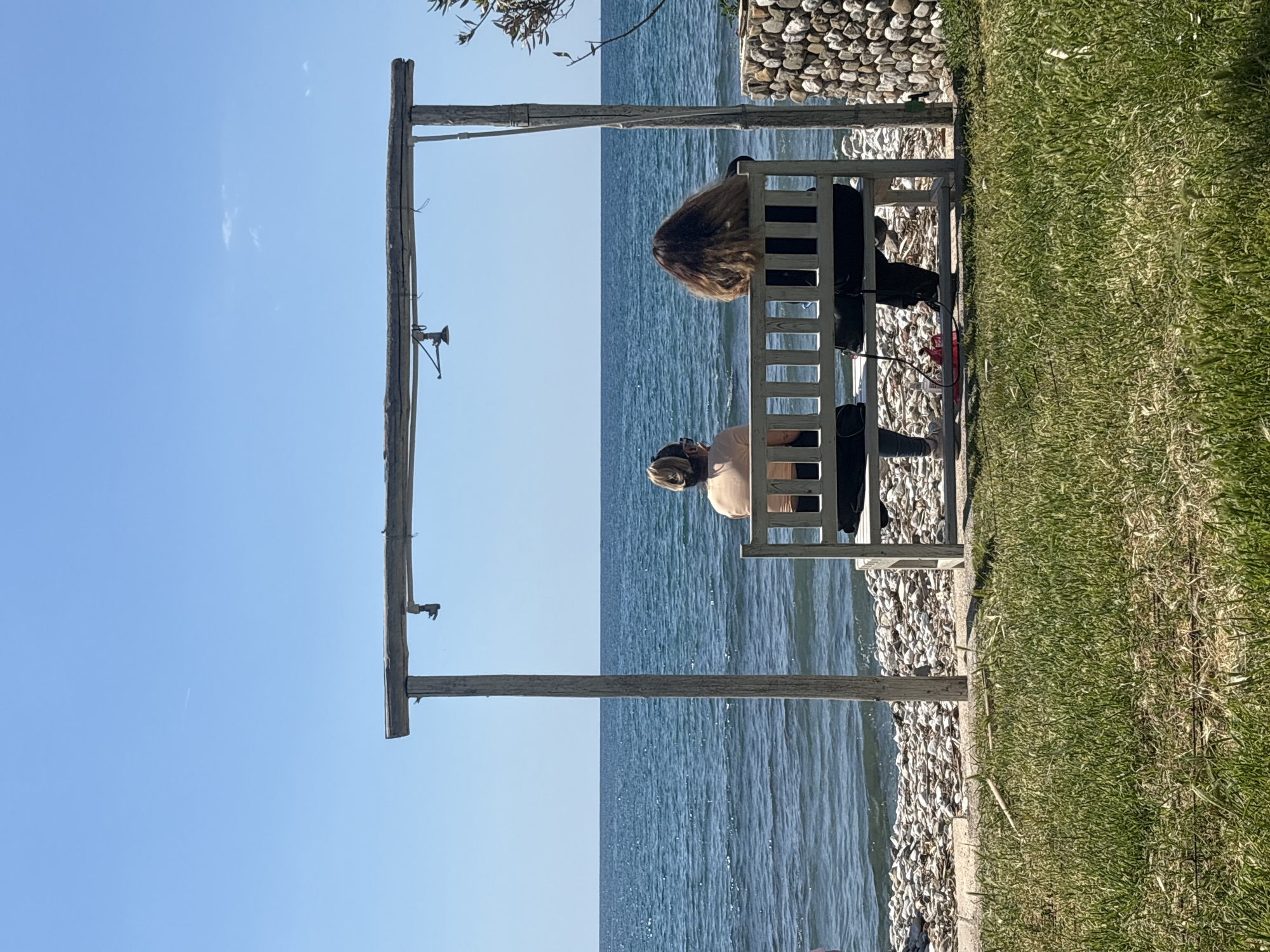 Two individuals are seated on a white, open-backed bench overlooking a body of water, potentially the Adriatic Sea. The scene is partially framed by a weathered wooden structure resembling a rudimentary gazebo or swing set frame. The coastline consists of pebbles and rocks, transitioning into a grassy area. The sky is clear and blue, indicating a sunny day. The location is likely in the city of Bar, Montenegro, considering the prompt's instructions.