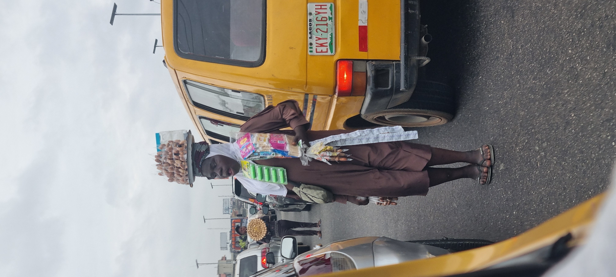 An outdoor medium shot captures a person standing on an asphalt road in what appears to be traffic, likely in Akowonjo, Nigeria. The individual, who is the central subject, wears a brown, loose-fitting garment and a white headscarf. They are carrying an assortment of goods. On their head, a flat, circular tray is balanced, stacked with numerous small, light-brown, round objects, possibly boiled eggs or nuts, enclosed in clear plastic. Attached to the front of their garment are several packages of brightly colored snacks and sweets, along with what appear to be long, thin white wrappers. A beige crossbody bag is slung over their left shoulder. The person is looking slightly to their right, with their mouth open. They are wearing flat, gray flip-flops.
To the right of the main subject is a yellow commercial van, a common sight in Lagos, with a license plate reading "EKY 216YH". The van has a black bumper and a red taillight. To the left of the main subject is a silver car, partially visible, with its rear taillight illuminated. Further in the background, other vehicles are present, indicating a traffic jam. The sky is overcast, with a diffused light. The overall scene depicts a vendor operating within the hustle and bustle of city traffic.