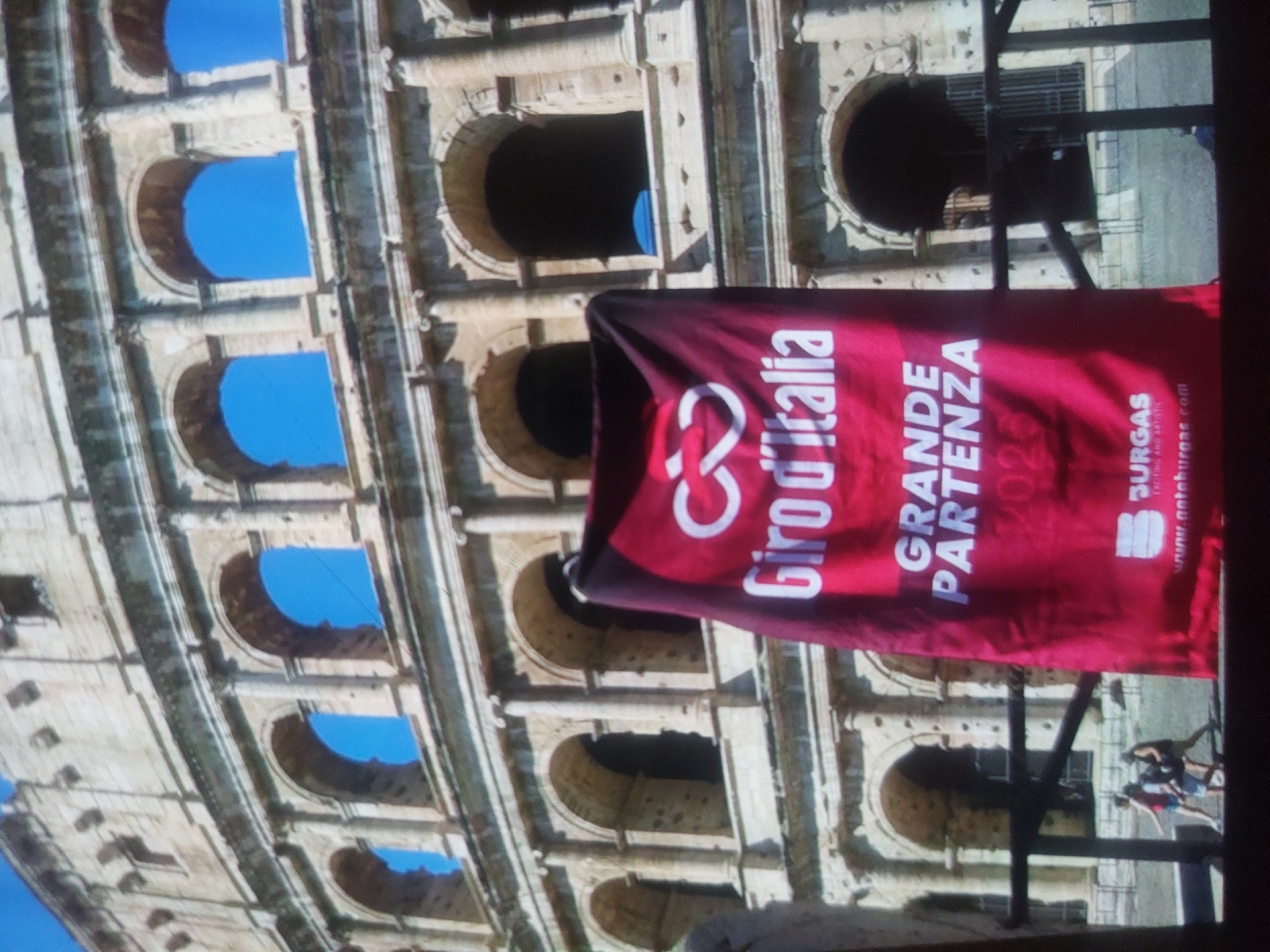 The image shows the Colosseum in Rome, Italy. A red banner advertising the Giro d'Italia Grande Partenza 2022 is prominently displayed. People are visible walking near the structure. There is no indication of Tsarevo, Bulgaria in the image.