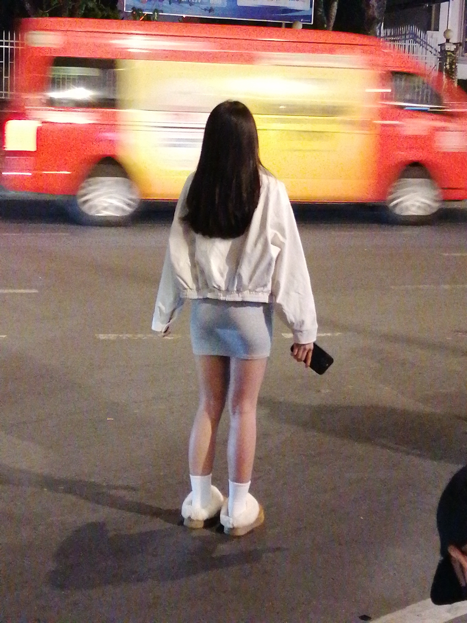 A young woman stands on a paved road at night, facing away from the camera. She has long, dark hair and is wearing a white cropped jacket over a light blue mini-dress. Her feet are clad in white socks and fluffy slip-on slippers. In her right hand, she holds a dark-colored smartphone.
Behind her, a red and yellow bus is in motion, appearing blurred due to its speed. The bus has white windows and its wheels are also indistinct. The road surface is asphalt with white lane markings visible. The scene appears to be a street in Da Lat, Vietnam, at night. The lighting suggests streetlights and ambient city illumination. The woman's posture is relaxed, and there are no discernible emotions conveyed. There is no visible text in the image.