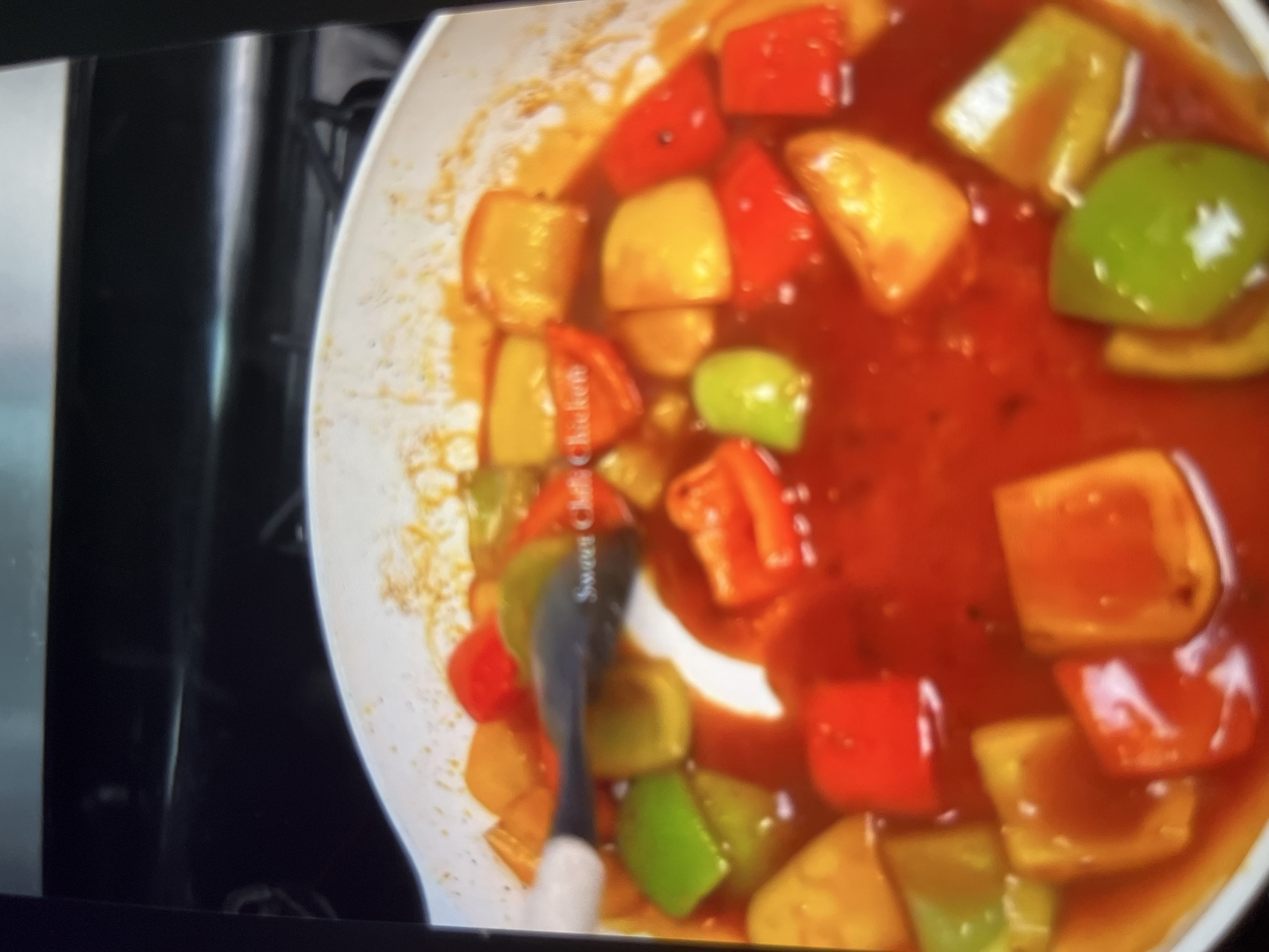 The image shows a close-up of a dish containing colorful pieces of bell peppers and possibly chicken in a thick, red sauce. A spatula with a white handle is partially visible, stirring the contents of what appears to be a white pan. The text "Sweet Chili Chicken" is faintly visible on the spatula. The background is dark and out of focus, suggesting a kitchen environment. There is no discernible information about the location in Abuja, Nigeria, as the image focuses solely on the food preparation.