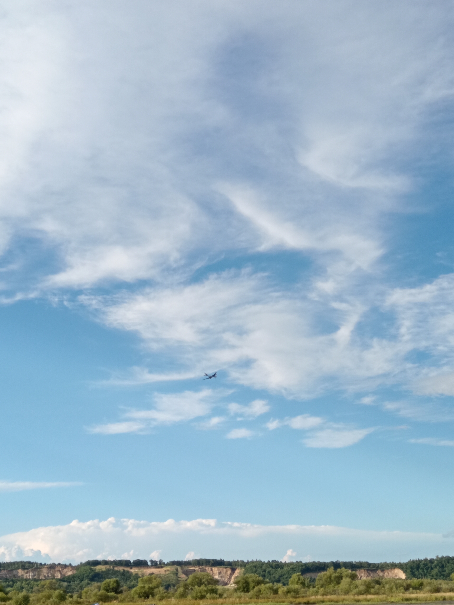 The image captures a wide expanse of a bright blue sky filled with wispy white clouds. A small airplane is visible flying in the upper right quadrant of the sky. Below the sky, a distant, rolling landscape is depicted. It features a strip of light-colored, eroded cliffs topped with dense green trees. A lighter green area of foliage stretches across the foreground. The scene appears to be outdoors, under a clear sky with scattered clouds, and suggests a natural or semi-rural environment.
