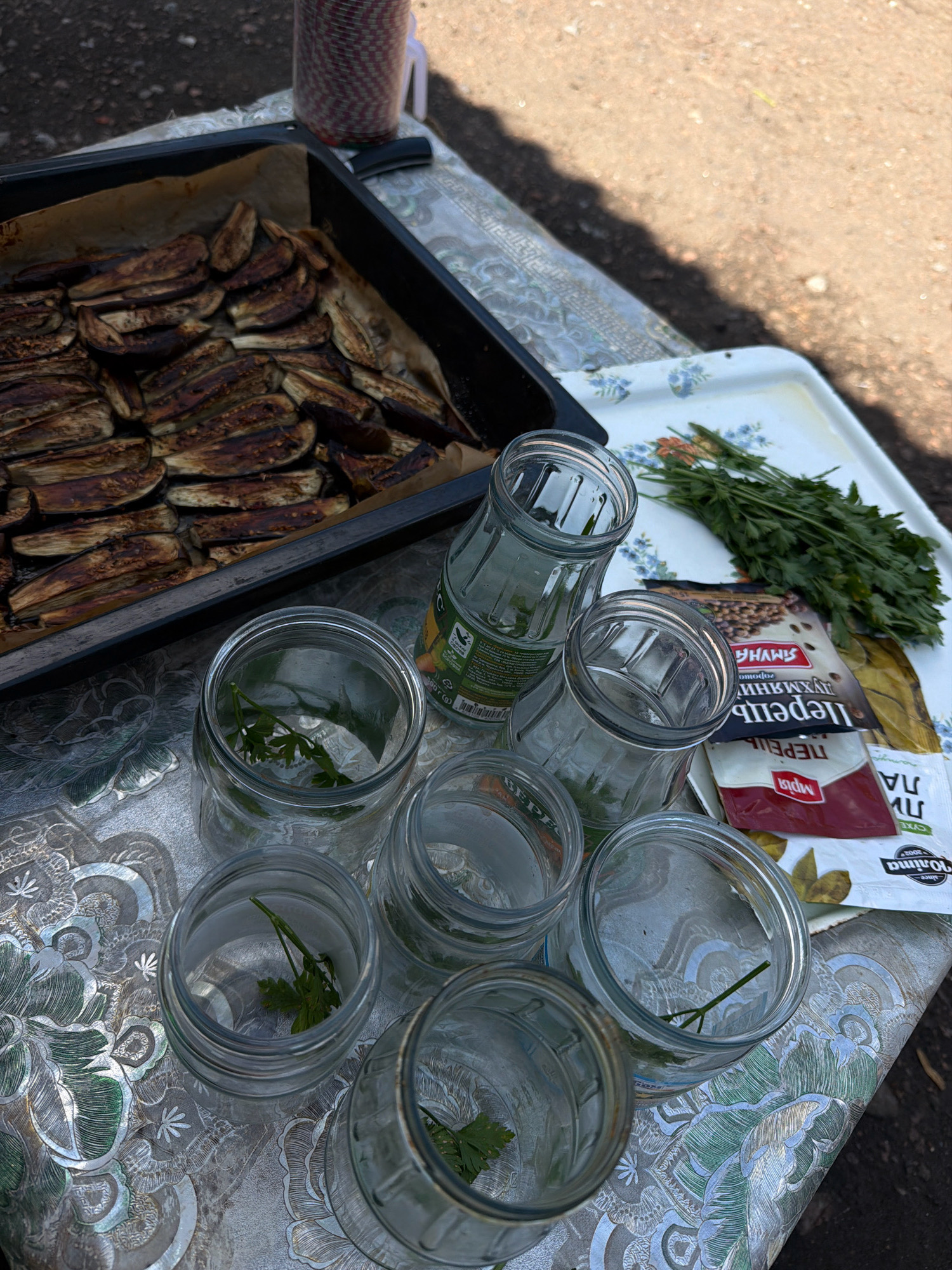 This is a high-angle, close-up shot of food preparation on a table outdoors, likely in Novosilka, Ukraine, given the context. In the foreground, several empty glass jars are arranged, some containing sprigs of parsley. Behind the jars, a white tray holds a bunch of fresh parsley and two small packets of spices, one red and white, the other brown and white with Cyrillic lettering. A bottle with a green label is also present. In the background, a baking tray is filled with roasted eggplant slices. The table is covered with a patterned tablecloth. The ground surface visible beyond the table appears to be gravel or dirt. The lighting suggests it is daytime with shadows cast on the scene.