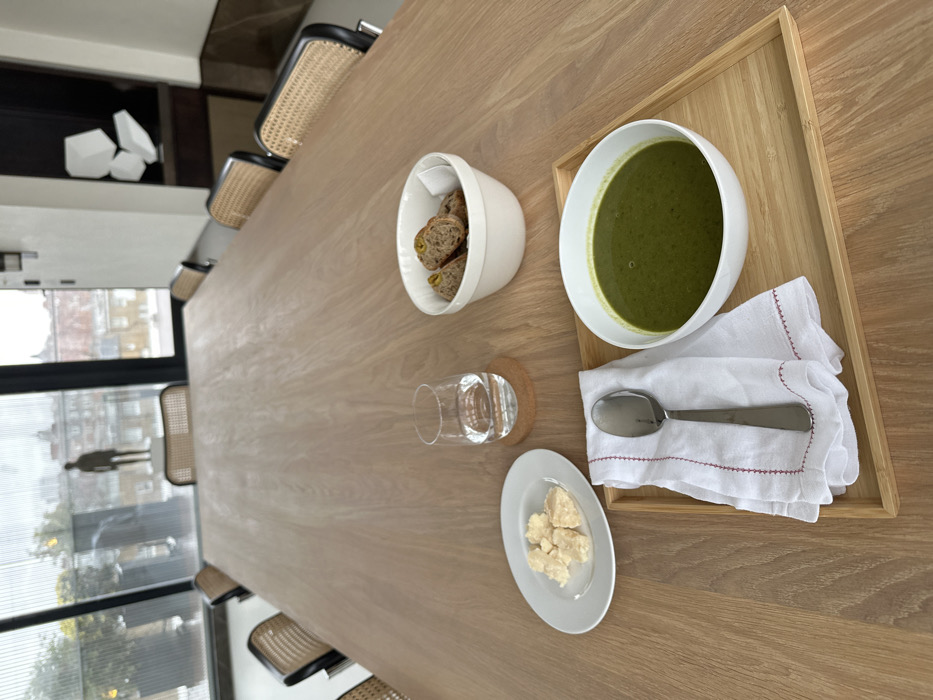 A formal meal setting is depicted on a long, light-toned wooden table within a modern, well-lit room, identified as The Conduit in Mayfair, London, United Kingdom. In the foreground, a light bamboo tray holds a white bowl of dark green soup, and a folded white linen napkin with red embroidered edges, atop which rests a silver spoon. To the tray's left, an oval white plate contains several irregular pieces of light-colored cheese. A clear glass of water on a cork coaster is positioned nearby. Further up the table, a white bowl holds multiple slices of dark bread. The room is furnished with chairs featuring black frames and woven cane backs along the table's length. A large window dominates the background, revealing an urban landscape of London rooftops and buildings under an overcast sky. A notable full-body sculpture of a standing male figure is visible on an outdoor terrace immediately beyond the window. Interior architectural elements include abstract white geometric wall art against a dark surface. No people are present.