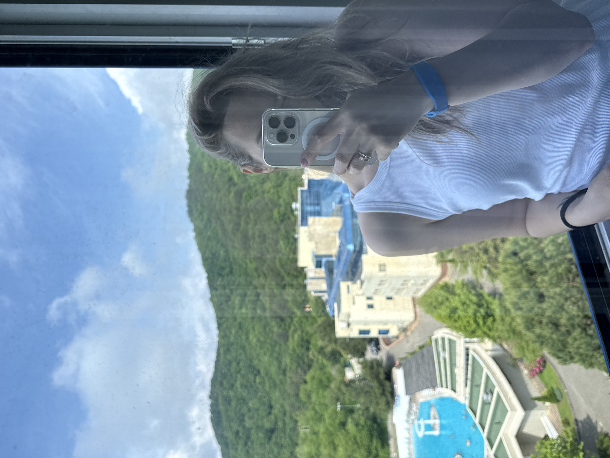 A woman is taking a selfie with her iPhone in front of a window overlooking a resort in Nebug, Russia. She is wearing a white tank top and a blue wristband. In the background, there is a swimming pool with people swimming, a multi-story building with a blue roof, and lush green mountains. The sky is blue with scattered clouds.