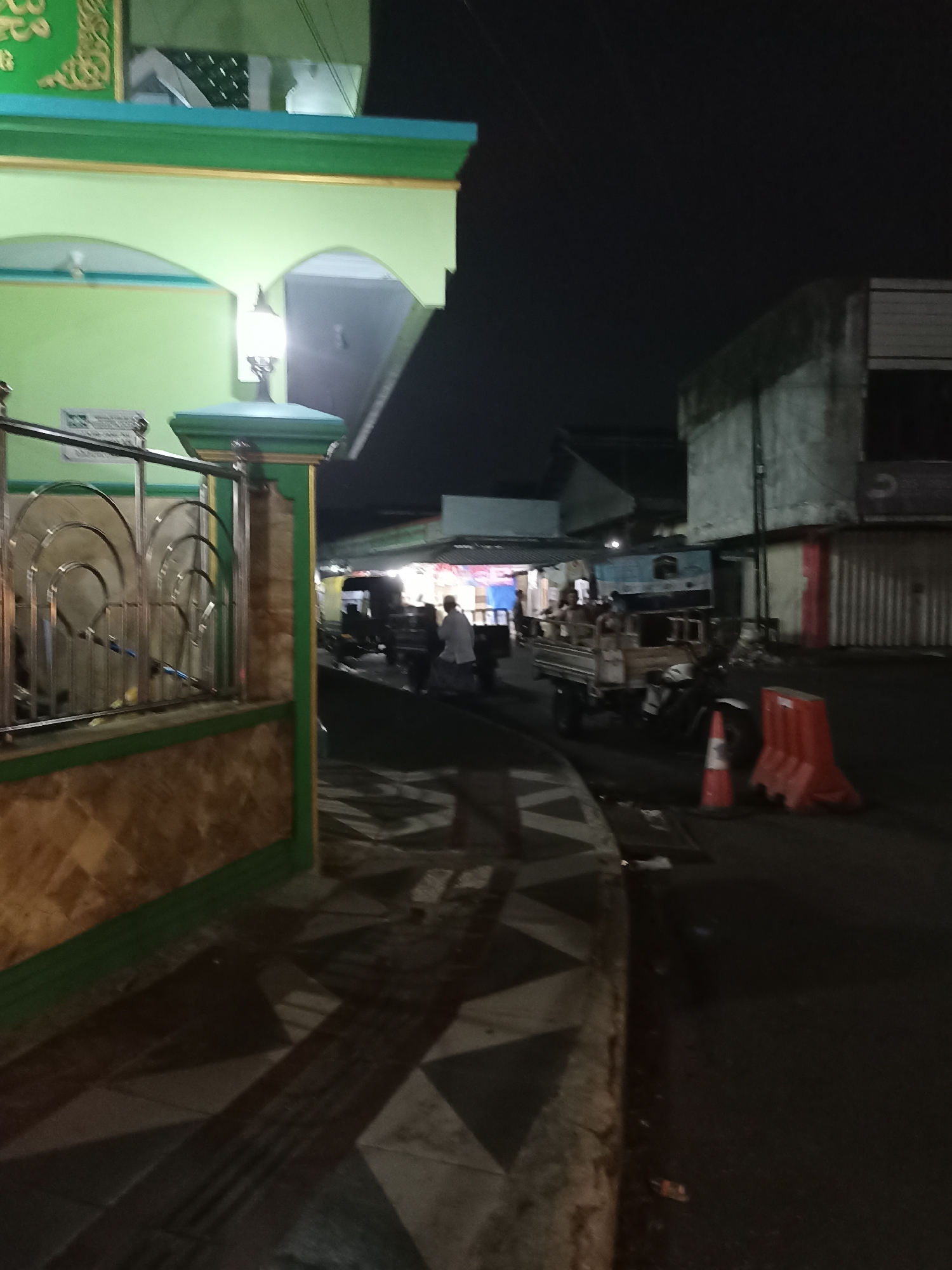 An exterior shot at night shows a dimly lit street in Tompokersari, Indonesia, with a building on the left and several structures on the right. The building on the left is light green with a decorative gold trim and a large archway, a lamp hangs from the archway. In front of the building is a stainless steel fence with a stone and light green base. The street in front of the building is paved with large tiles. On the right side of the street, there is a three-wheeled truck carrying people, and in the foreground, there are two orange traffic cones and two orange barriers. Several people are visible in the street, some near the truck and others walking in the background. The overall scene depicts a street in an urban area at night.