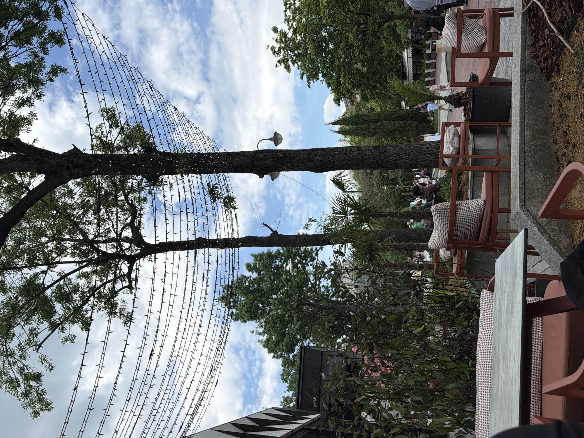 The image shows an outdoor seating area, possibly a restaurant or café, in what is likely the city of Varna, Bulgaria, given the mention in the prompt. The scene is viewed from a low angle, looking upwards and somewhat tilted.
Two prominent trees stand in the foreground and middle ground. Strands of small lights are hung between them, creating a festive atmosphere. The lights are unlit. One of the trees has a light fixture attached to its trunk.
To the right of the trees is a seating area with several chairs with patterned cushions and a table partially visible in the foreground. Behind the chairs is a raised platform or step, and in the background, there are people walking and seated in an outdoor public area. The greenery includes taller trees, shrubs, and some palm trees.
The sky above features a mix of blue patches and white clouds.