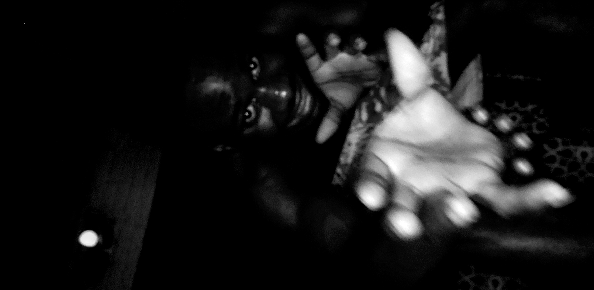 A striking, low-light, black-and-white portrait captures a person in Narayi, Nigeria. The individual, seen from an upward angle, appears in the upper-left and center of the frame, looking directly at the viewer with an intense, captivating gaze. Their eyes are clearly visible and reflective, contrasting with the deep shadows that obscure much of their face and head.
Two hands are prominently extended towards the camera, creating a dramatic, wide-angle effect. The hand in the foreground is large, slightly out of focus, and stretches out with fingers partially spread. The second hand is positioned behind it, also reaching forward. The background is largely enveloped in darkness, though a faint, circular, geometric pattern is discernible on a fabric to the right, possibly clothing or upholstery. A small, bright, out-of-focus light source appears in the far lower-left corner. The overall mood is intimate and mysterious, characterized by strong contrasts and a grainy texture indicative of low-light photography. No text is visible in the image.