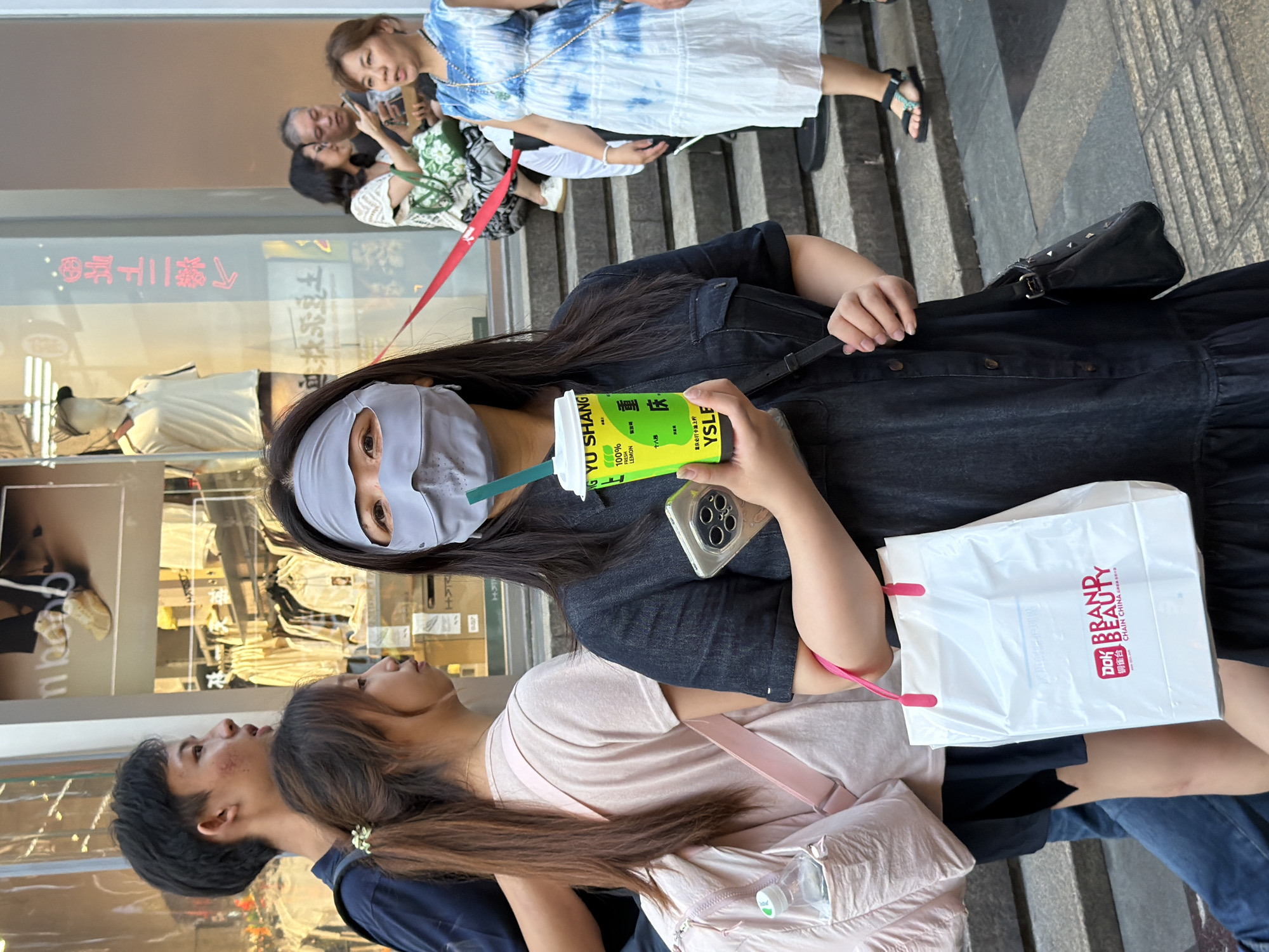 This image depicts an outdoor scene, likely a street in Yuzhong District, China, given the context. A young woman in the foreground, wearing a dark denim dress and a gray face covering that extends over her eyes and nose, holds a bright yellow beverage cup with a green straw. She is also carrying a white shopping bag with pink handles, which has "BRAND BEAUTY" and "CHAIN CHINA" printed on it in red text. To her left, a young man in a dark blue t-shirt and jeans is partially visible, and to his left, a young woman with her hair in a ponytail is seen from the side. In the background, a store window displays clothing and mannequins. Other individuals are visible further in the background, including a woman in a tie-dye dress and people sitting on steps. The ground appears to be paved with stone tiles.