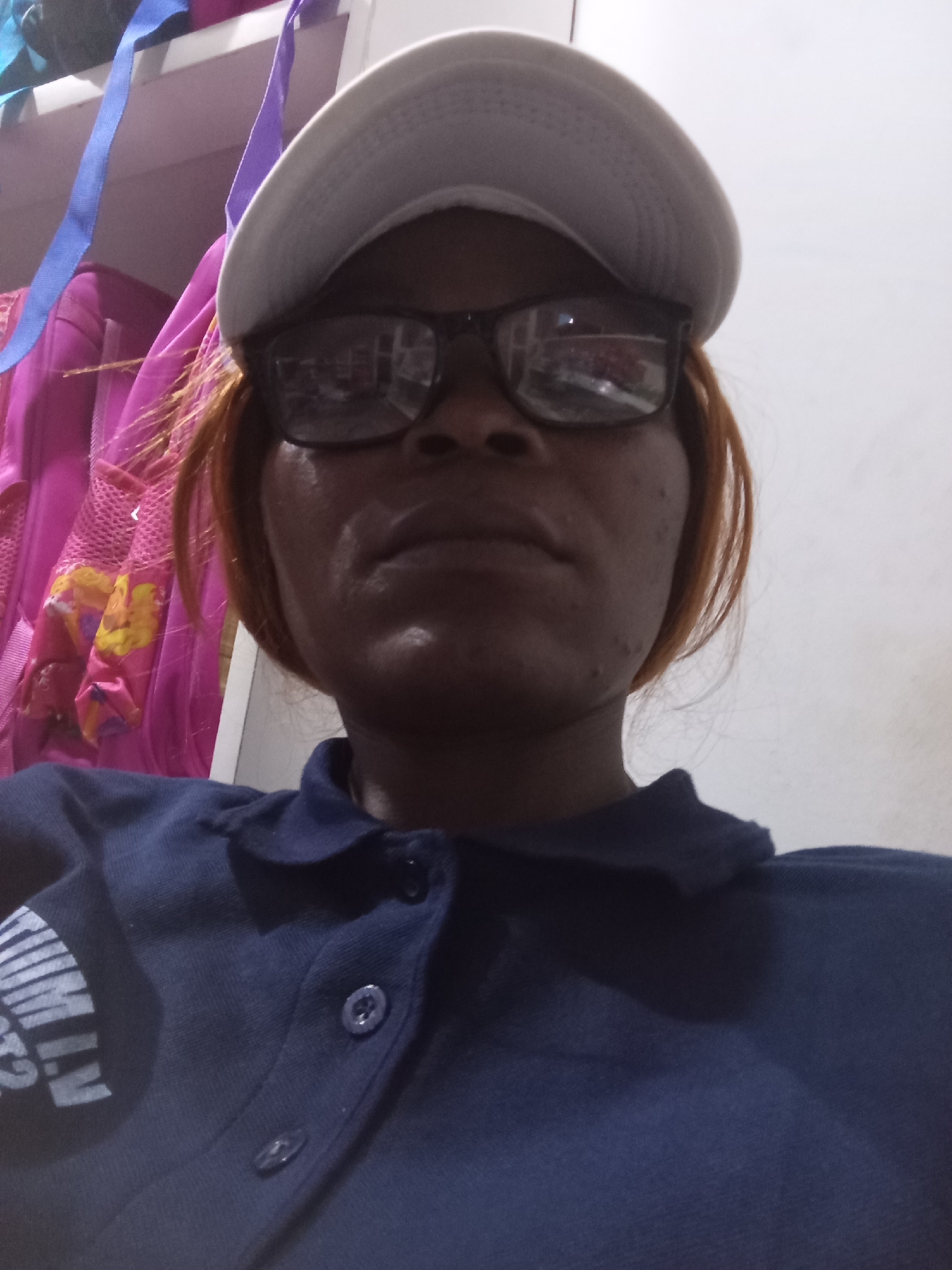 A close-up, low-angle selfie of a woman wearing a dark blue polo shirt and a white baseball cap. She has reddish-brown hair, dark-rimmed glasses, and her skin has a warm tone with some visible texture. The background is out of focus, showing a white wall and what appears to be a shelf with colorful items, possibly backpacks or bags, including a bright pink one with yellow accents, and some blue and purple straps hanging down. The scene is indoors, likely a residential setting in Howayi, Nigeria.