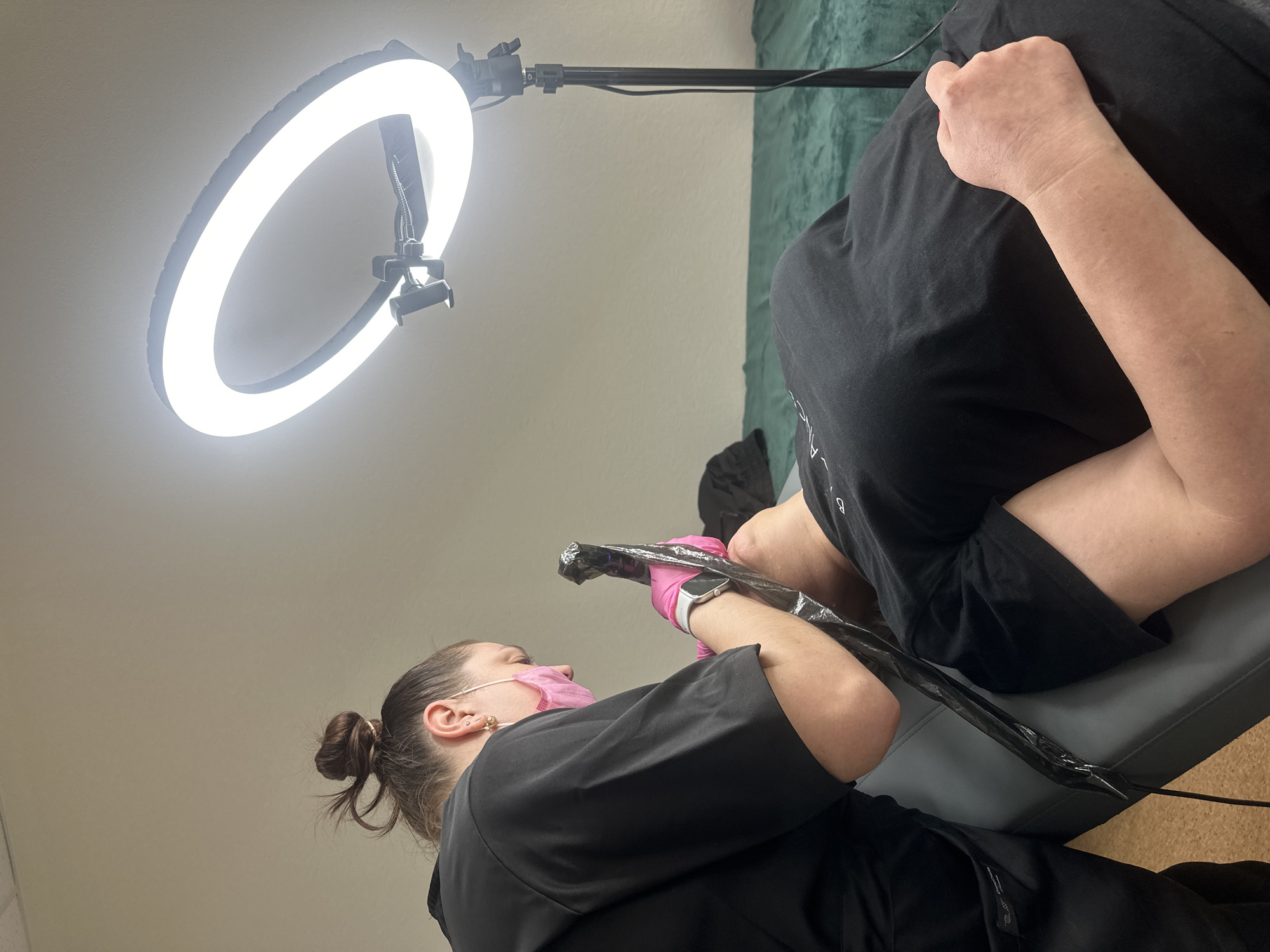A person wearing a black shirt and a pink face mask is tattooing another person's leg. The tattoo artist is wearing pink gloves and holding a tattoo gun. The leg being tattooed is covered in black plastic wrap. A ring light is illuminating the scene. The setting appears to be a tattoo parlor. The location is unknown, as there is no information provided in the image to indicate it is Chaikovsky, Russia.