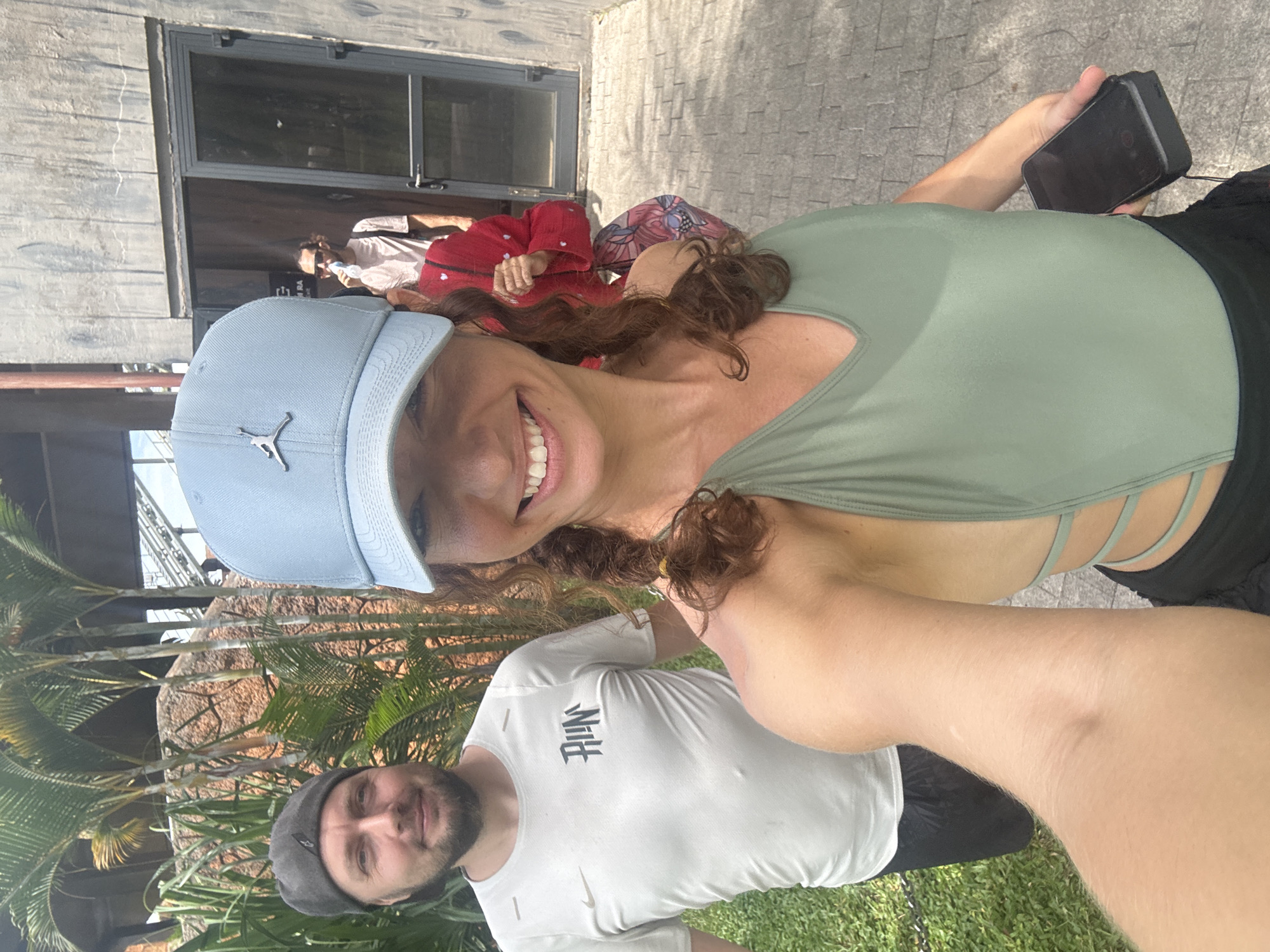 A selfie photograph taken outdoors in Đà Nẵng, Vietnam features a light-skinned woman wearing a light blue baseball cap and a green top, smiling at the camera while holding a mobile phone. Beside her, a light-skinned man with a beard is wearing a gray hat and a white Nike t-shirt. They are positioned near a building with a glass window and stone exterior, with palm trees visible in the foreground. In the background, other individuals are seated in what appears to be an outdoor dining area. The ground consists of grass and stone tiling.