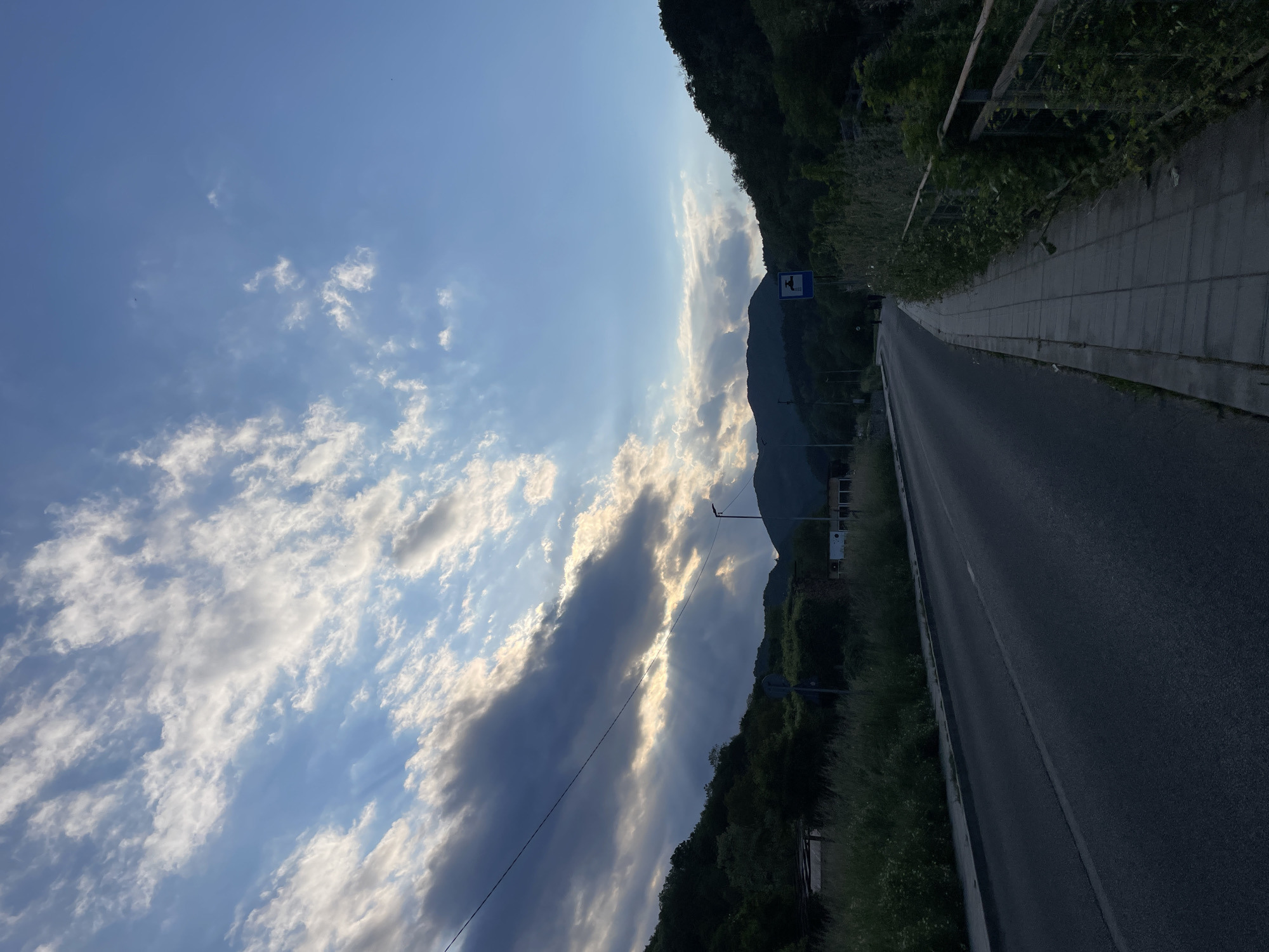 The image shows a road in Zlatograd, Bulgaria, flanked by a sidewalk on the right and a forested hillside on the left. The road extends into the distance towards a mountain. The sky is blue with scattered white and gray clouds, through which sunlight streams. A road sign is visible on the right side of the road, near the hillside. A metal guardrail is along the edge of the sidewalk.