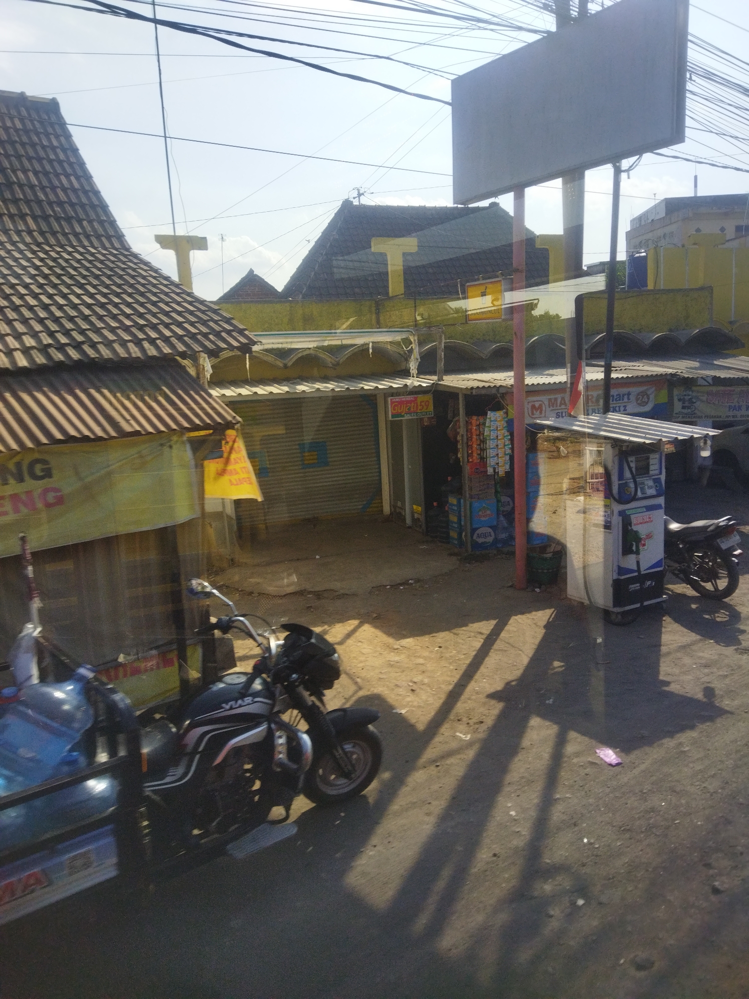 The image depicts a roadside scene in Nguter, Indonesia, featuring a small shop with a gas pump and parked motorcycles. In the foreground, a black motorcycle is parked on the dirt road, casting a shadow. To the left of the motorcycle, a blue cart with large plastic water bottles is attached to a different, partially visible motorcycle. The shop itself is made of corrugated metal and wood, with a peaked roof. A sign with yellow and red text is visible on the left side. A small, white and blue gas pump unit stands in front of the shop, also casting a shadow. Behind the shop, more structures and signage are visible, including a "Maret Mart 24" sign. The sky is bright blue with a few clouds, and electrical wires crisscross overhead. The ground is a mixture of dirt and gravel, with scattered debris. The scene appears to be a small, local business operating along a street.