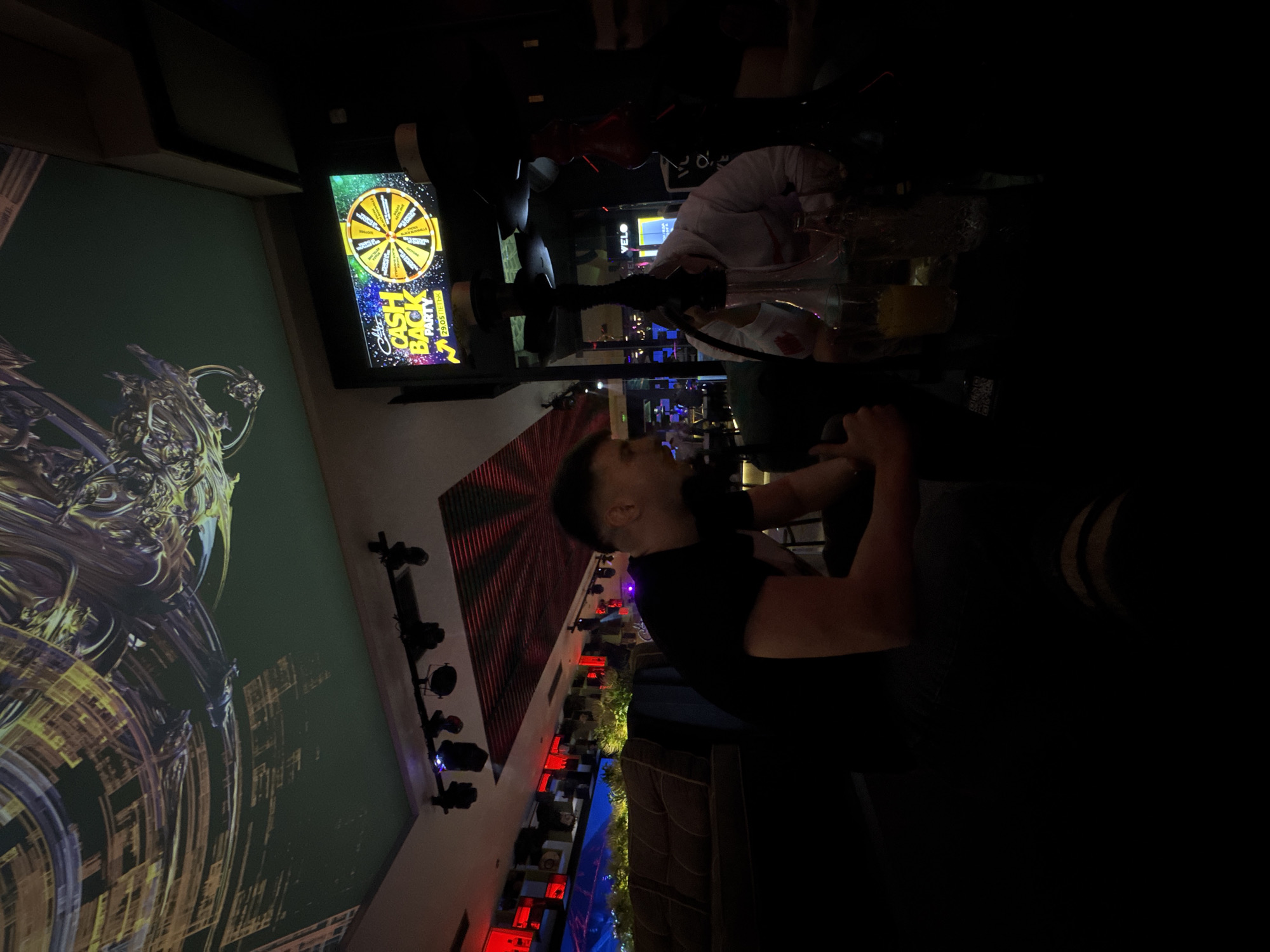 The image shows the interior of a nightclub or bar in Sofia, Bulgaria. A man in a black t-shirt sits at a table, possibly smoking a hookah. In the background, there is a woman wearing a white shirt. A promotional screen showing "Cash Back Party 29/05" is displayed. The club features modern decor with illuminated walls and lighting.