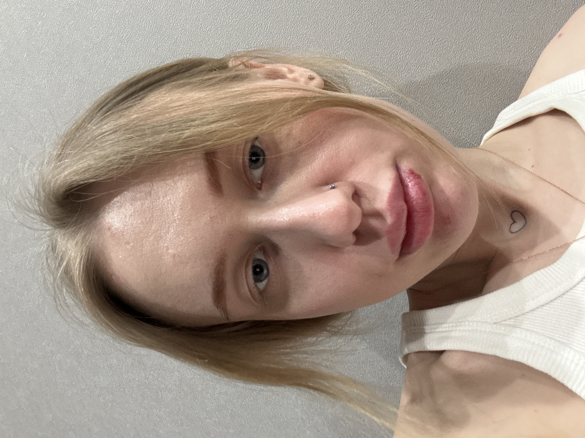 A close-up, head-and-shoulders portrait depicts a fair-skinned individual, appearing female, with blonde hair and blue-grey eyes, facing directly towards the viewer. A small, dark stud piercing is present on the individual's left nostril. The lower lip exhibits a visible lesion or injury on the left side, appearing red and partially crusted. The individual is wearing a white ribbed tank top. A thin, gold-colored necklace is visible, along with a small, black outline heart tattoo on the left side of the neck, above the clavicle. The background is a plain, light-grey textured wall, consistent with an indoor environment. The image contains no visual information to determine the city of Rostov-on-Don, Russia, or any specific location within it.
