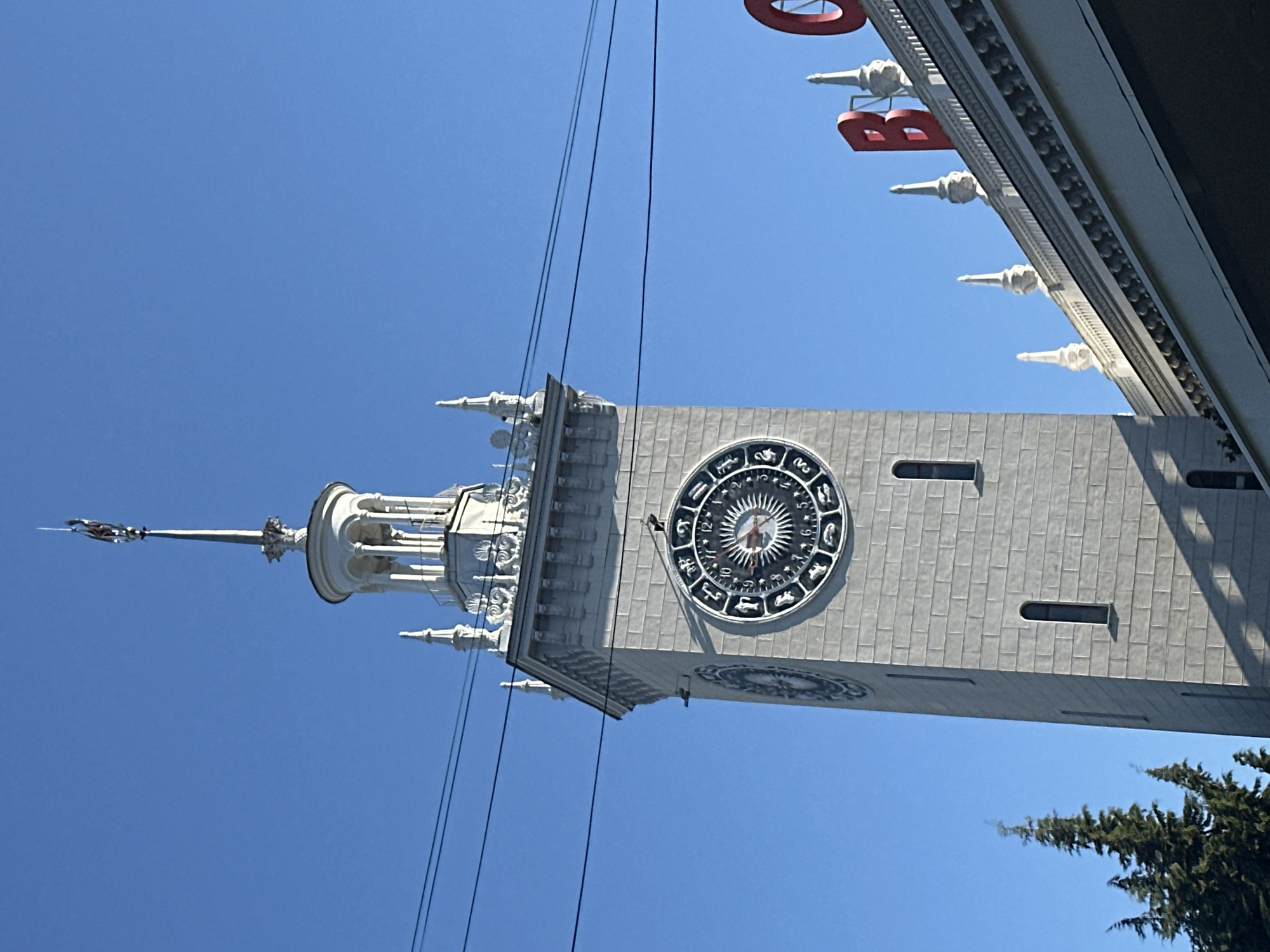 The image shows the clock tower of the Sochi railway station, a prominent landmark in the city of Sochi, Russia. The clock tower is a tall, grey, brick building with a white, ornate top section featuring a spire. The clock face on the tower is circular and displays zodiac signs around its perimeter, with a sun-like design in the center and hour and minute hands. To the right of the clock tower, part of a building with red lettering spelling out "ВОКЗАЛ" (station) is visible, as well as decorative white spires along the roofline. The sky is a clear, bright blue, and power lines are strung across the upper part of the frame. In the lower left corner, the tops of dark green evergreen trees are visible. The scene is bathed in sunlight, casting shadows on the clock tower and the building to the right. There are no people visible in the image.
